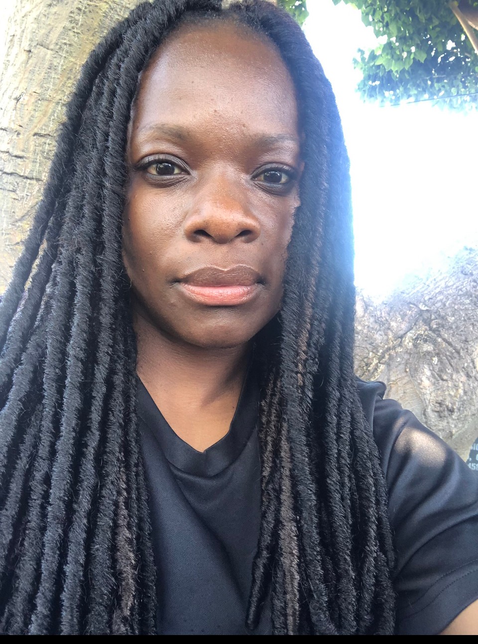 Woman with long dreadlocks outdoors, standing near a tree, looking at the camera, sunlight on her face.