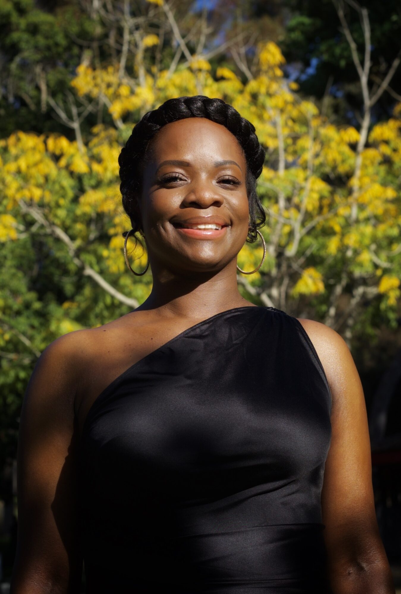 Smiling woman in black dress outdoors with yellow flowering tree in background.