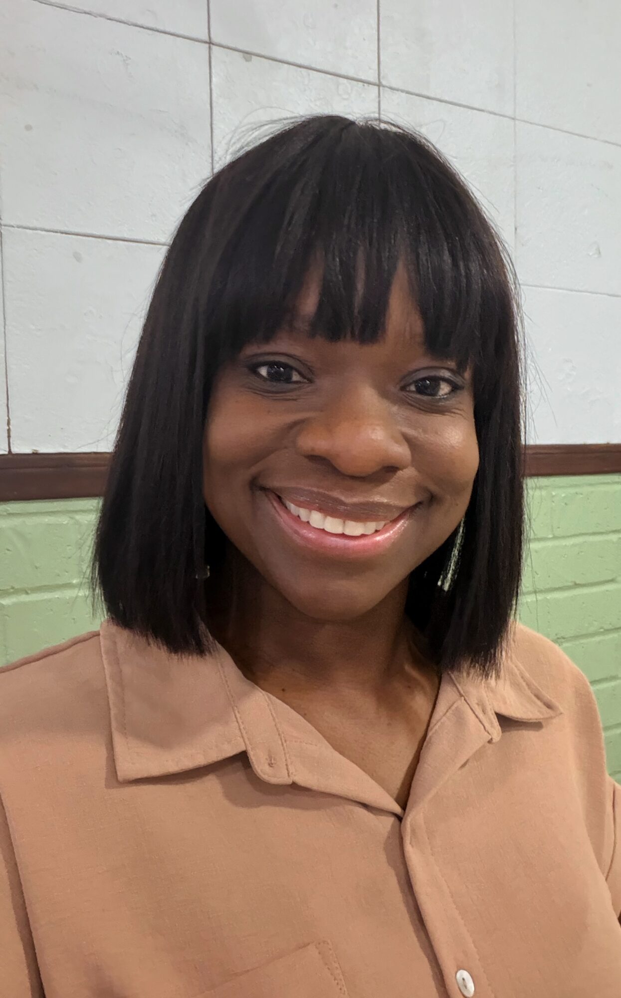 Smiling woman with shoulder-length black hair and bangs, wearing a beige collared shirt, standing indoors.