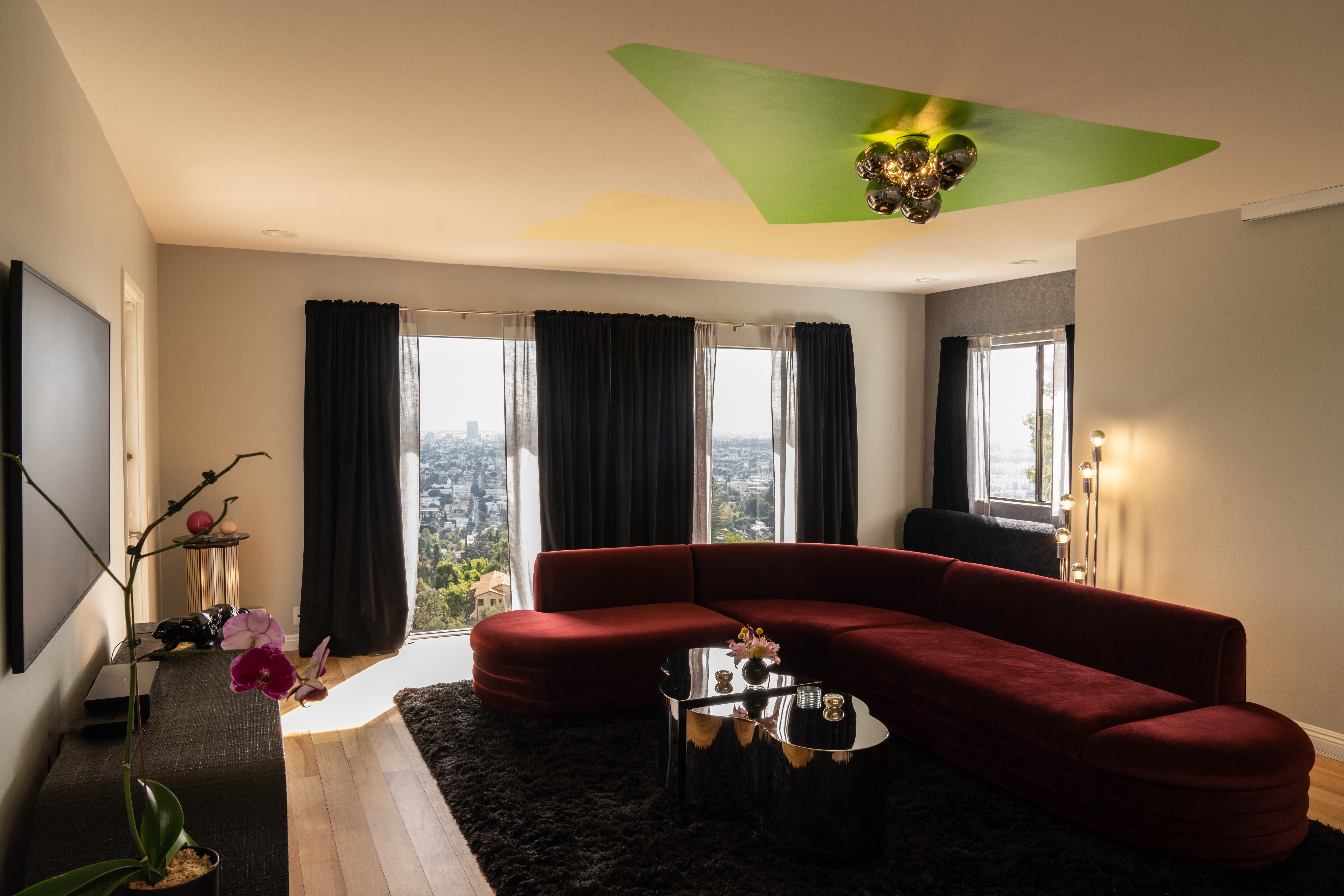 Living room with large windows, black curtains, red curved sofa, black rug, and a ceiling with green and yellow design.