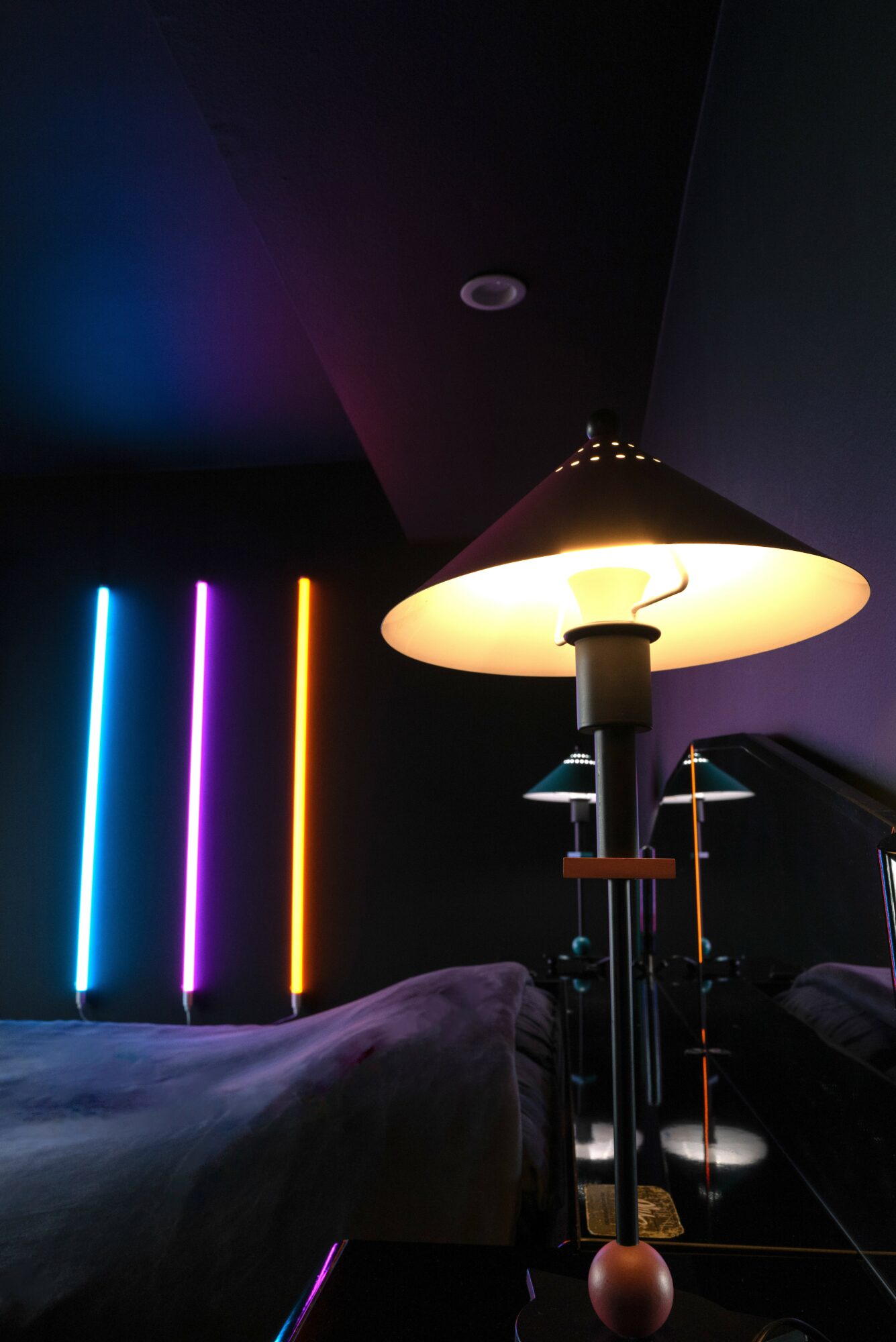 Room with colorful vertical neon lights, a large lamp, and a bed with dark bedding, ceiling and wall visible.