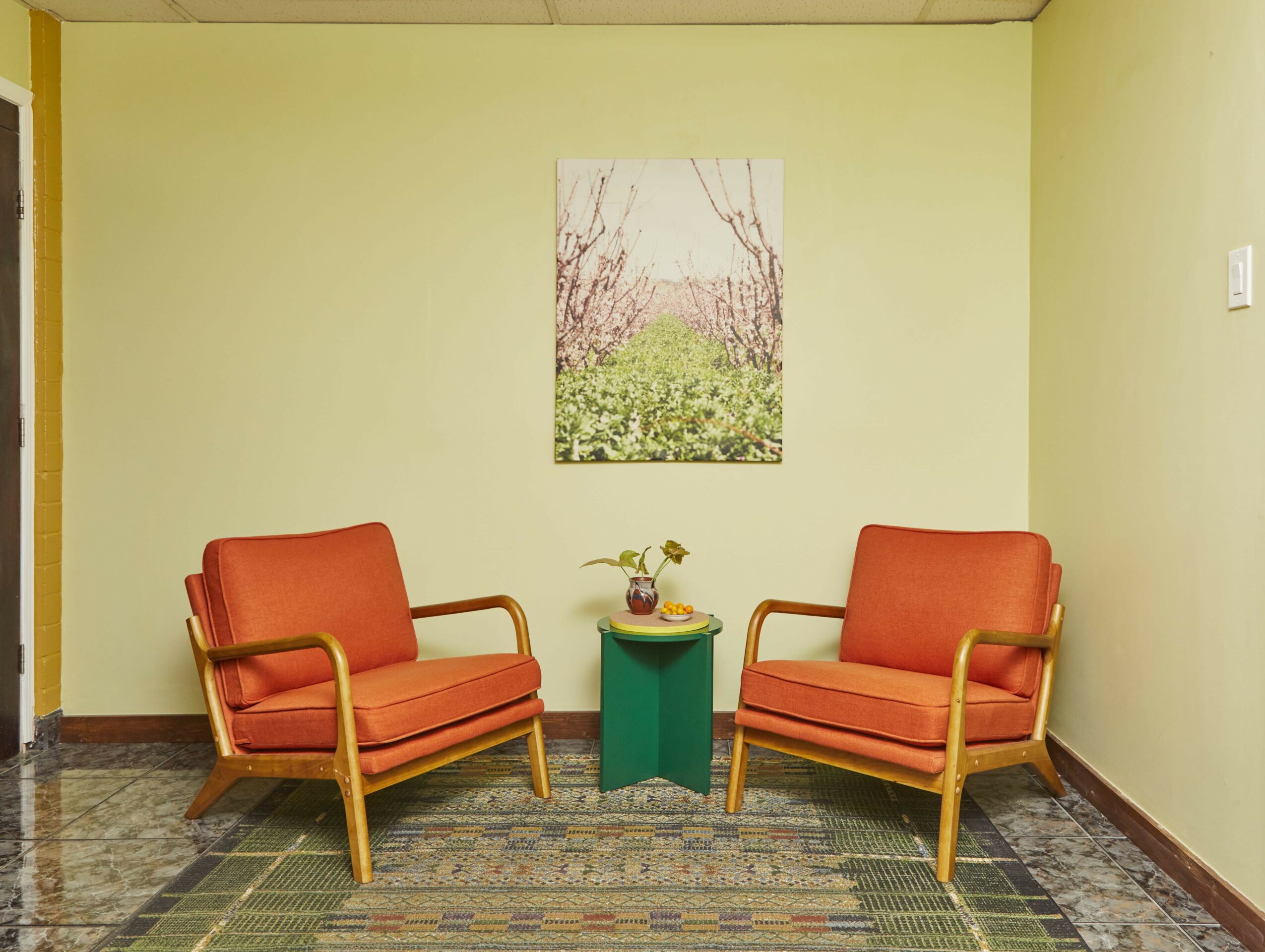 Two orange chairs with wooden arms and legs face each other with a small green table between them, a painting hangs on a light green wall.