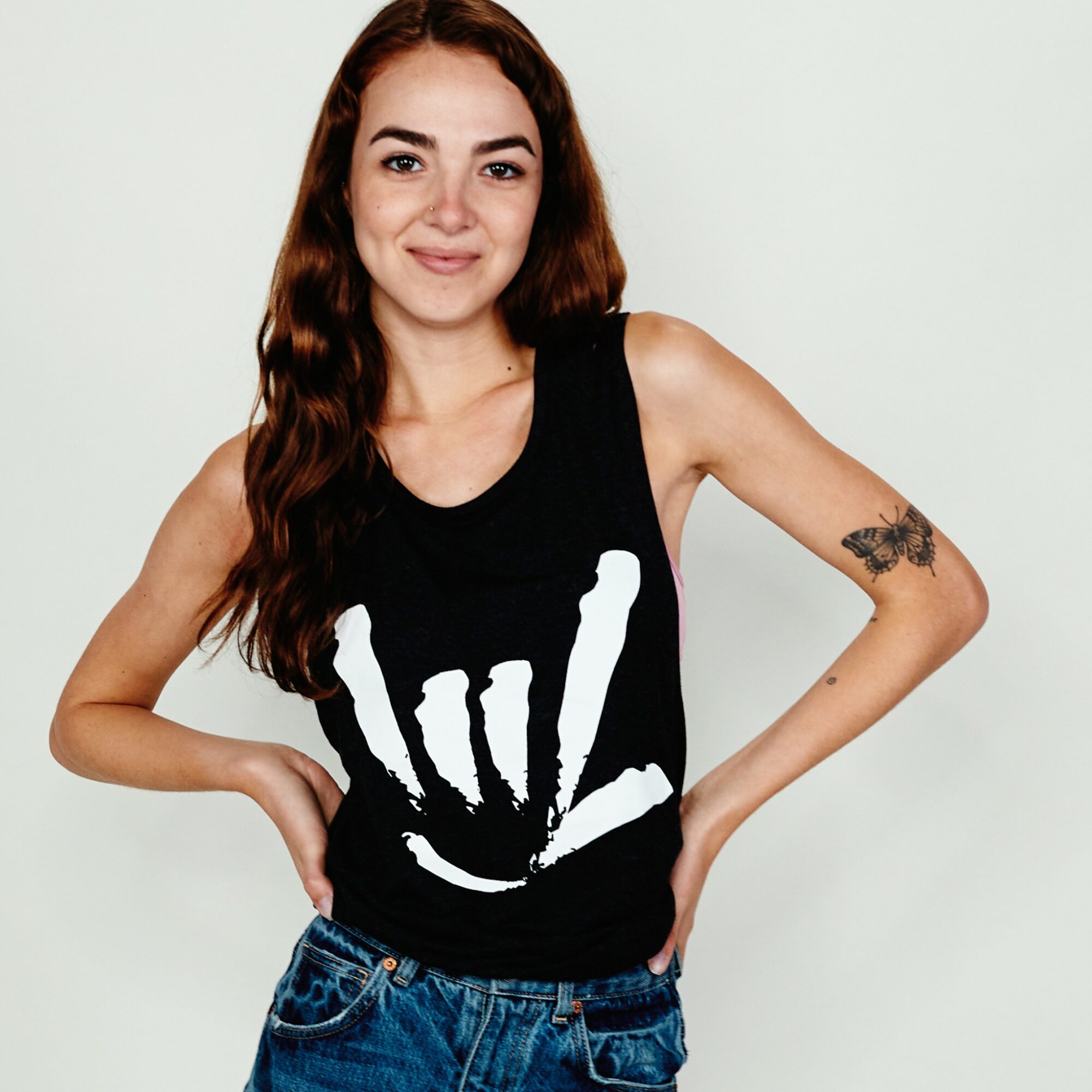 Young woman with long wavy hair wearing a black sleeveless top with a white handprint design, smiling, standing with hands on hips.