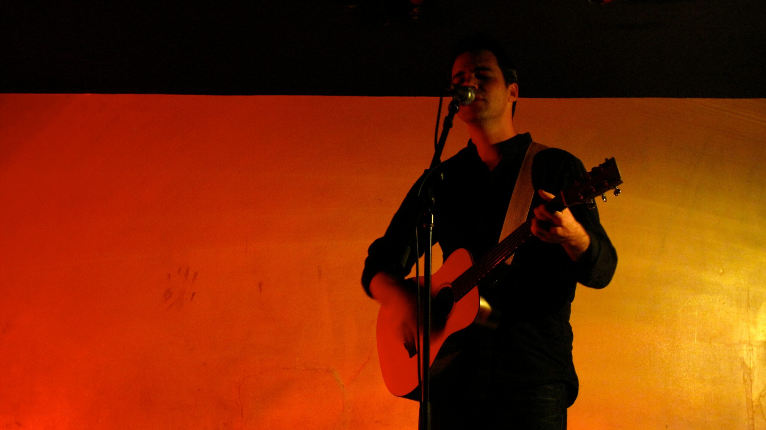 Person playing guitar and singing into microphone on stage with orange background.