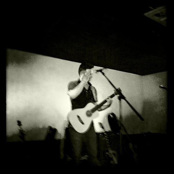 Person with guitar singing into microphone on stage, with a plain background and some equipment visible.