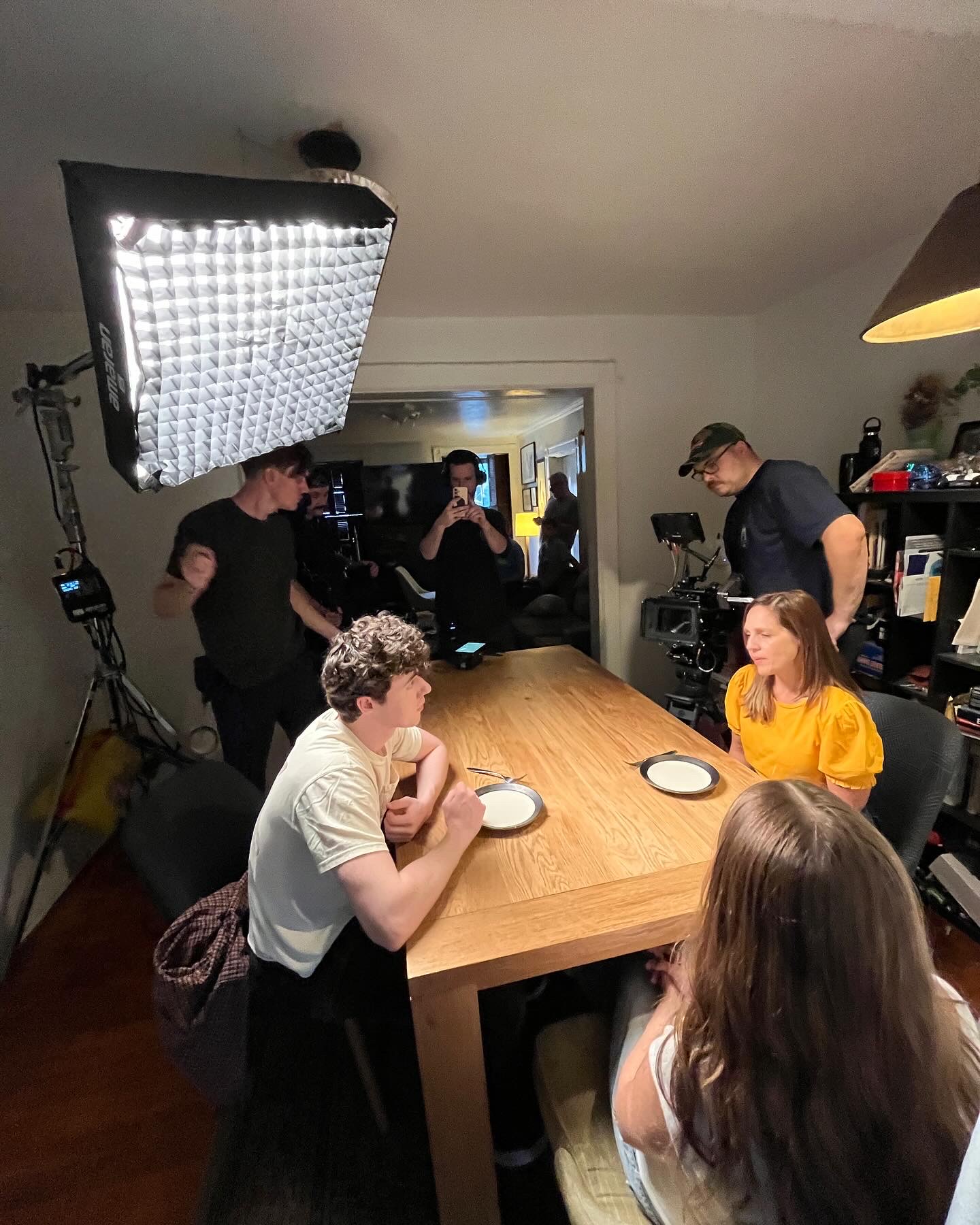 People gathered around a wooden table in a room with filming equipment, including a large light and camera, during a filming setup.