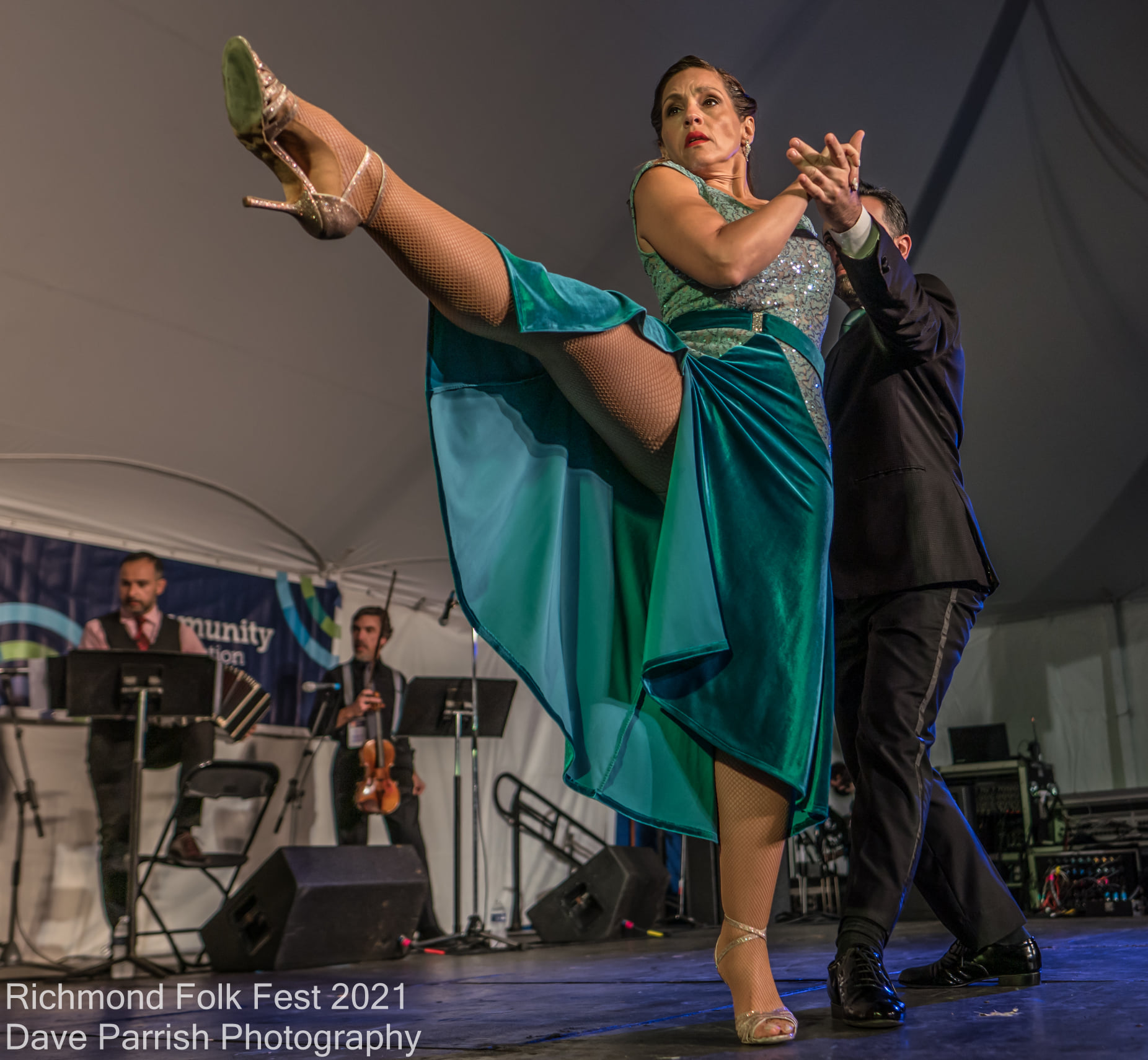 A woman in a teal dress performs a dance move with a raised leg, on stage with musicians in the background.