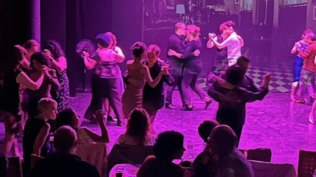 People dancing and socializing in a dimly lit room with purple lighting, some seated in the foreground.