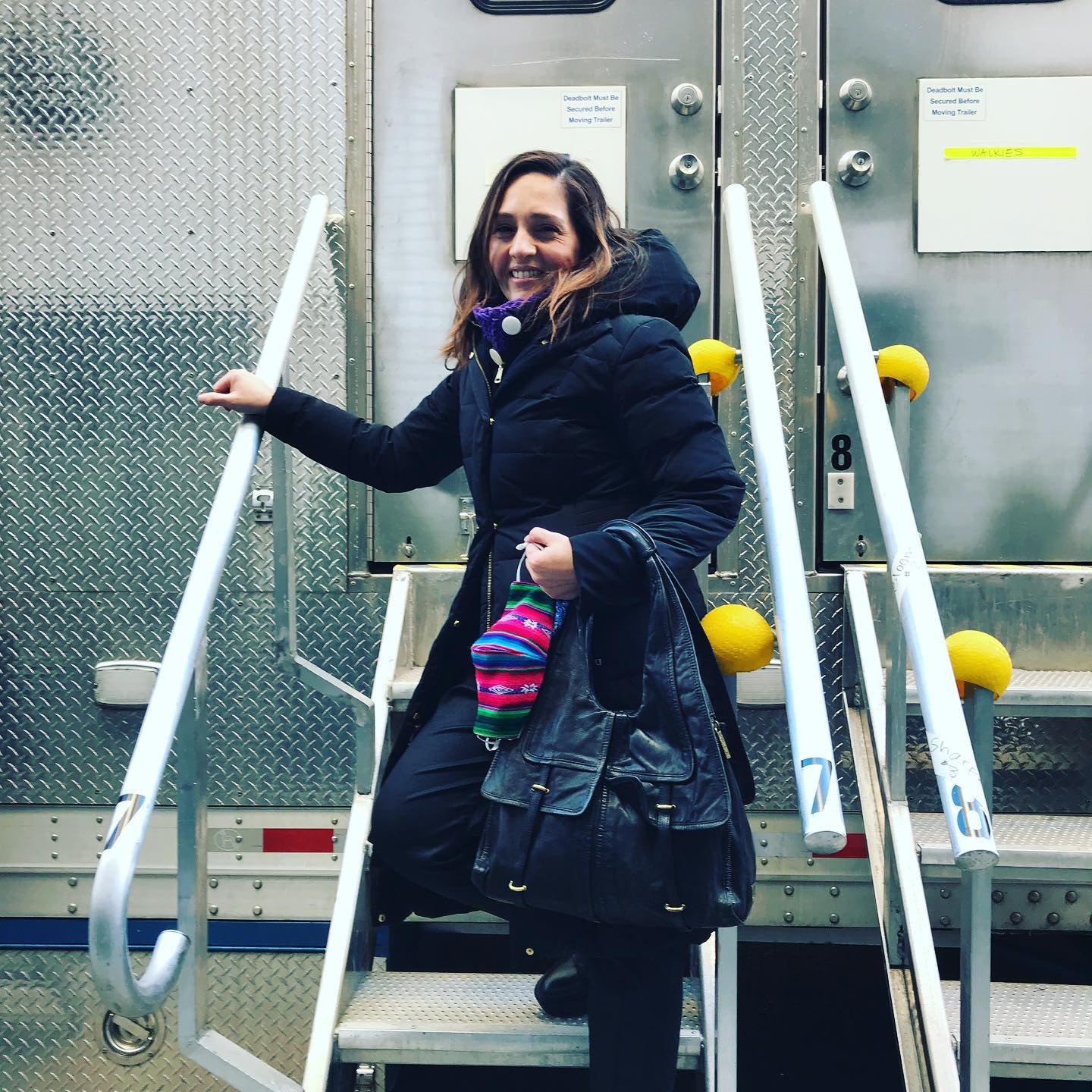 Woman smiling, sitting on stairs outside a metallic building, wearing a dark coat and colorful gloves, holding a black bag.