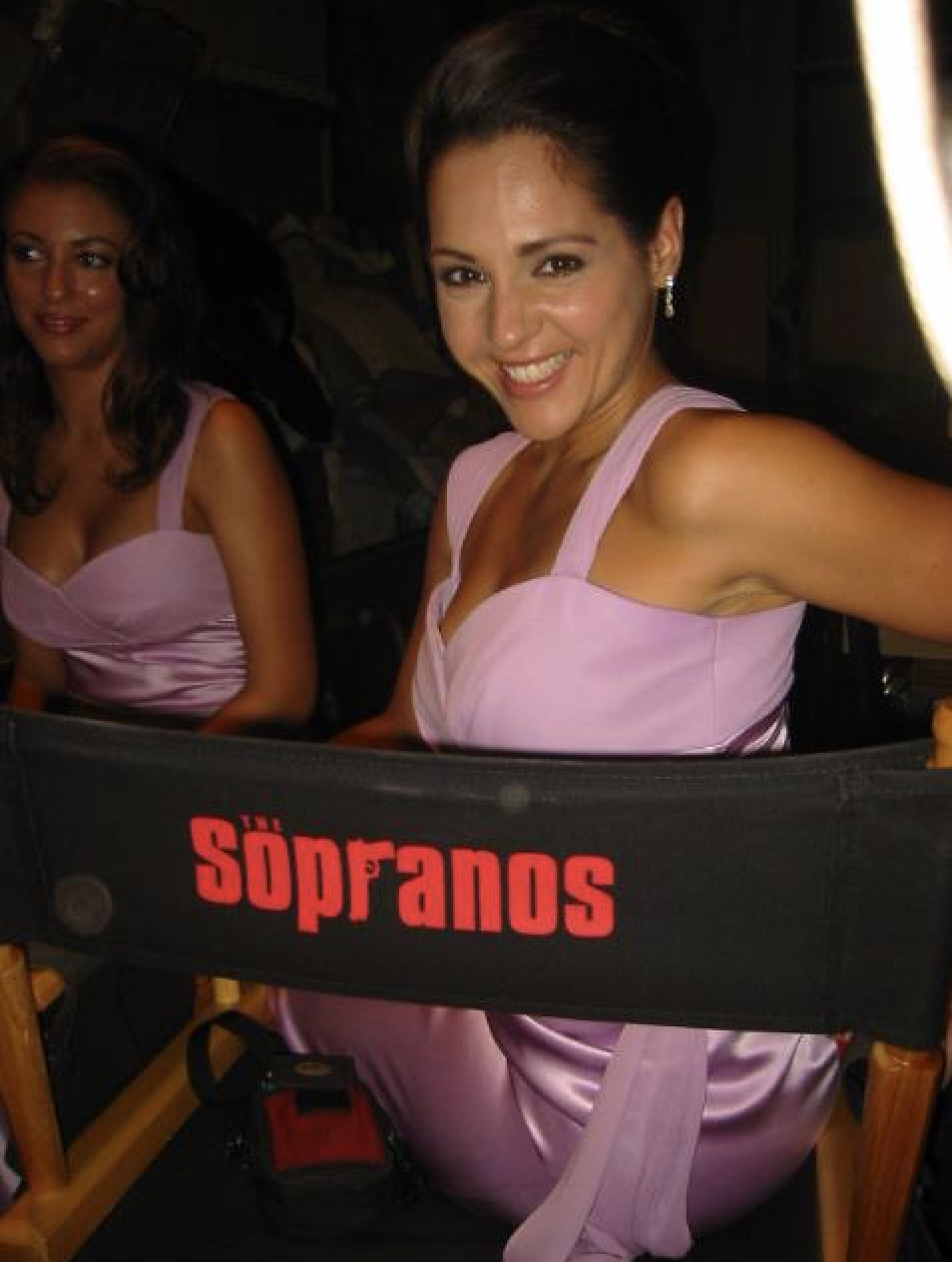 Two women in pink dresses sitting behind a black barrier with red text, smiling at an event.