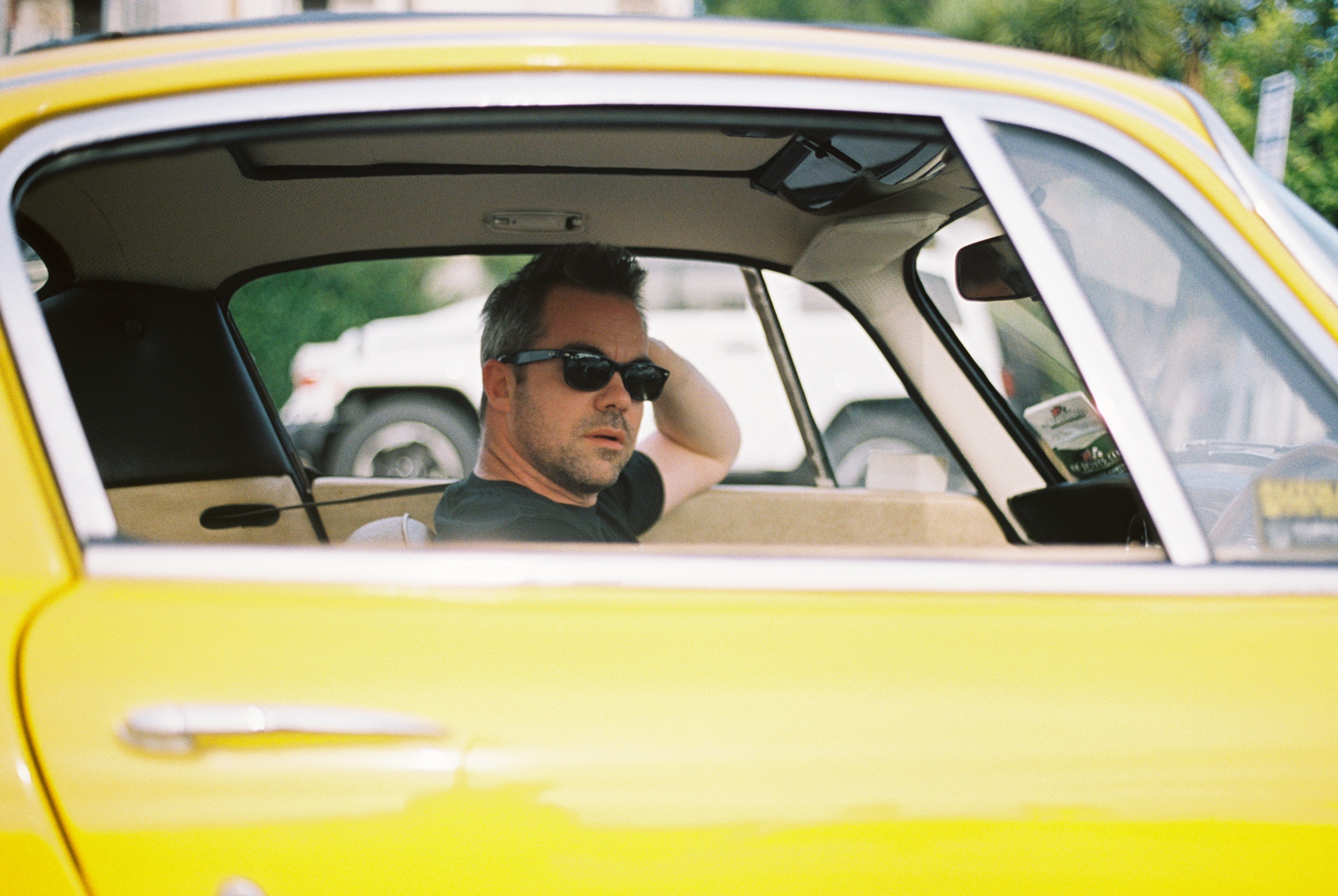 Man wearing sunglasses sitting in a yellow car, looking out the window with one hand behind his head.