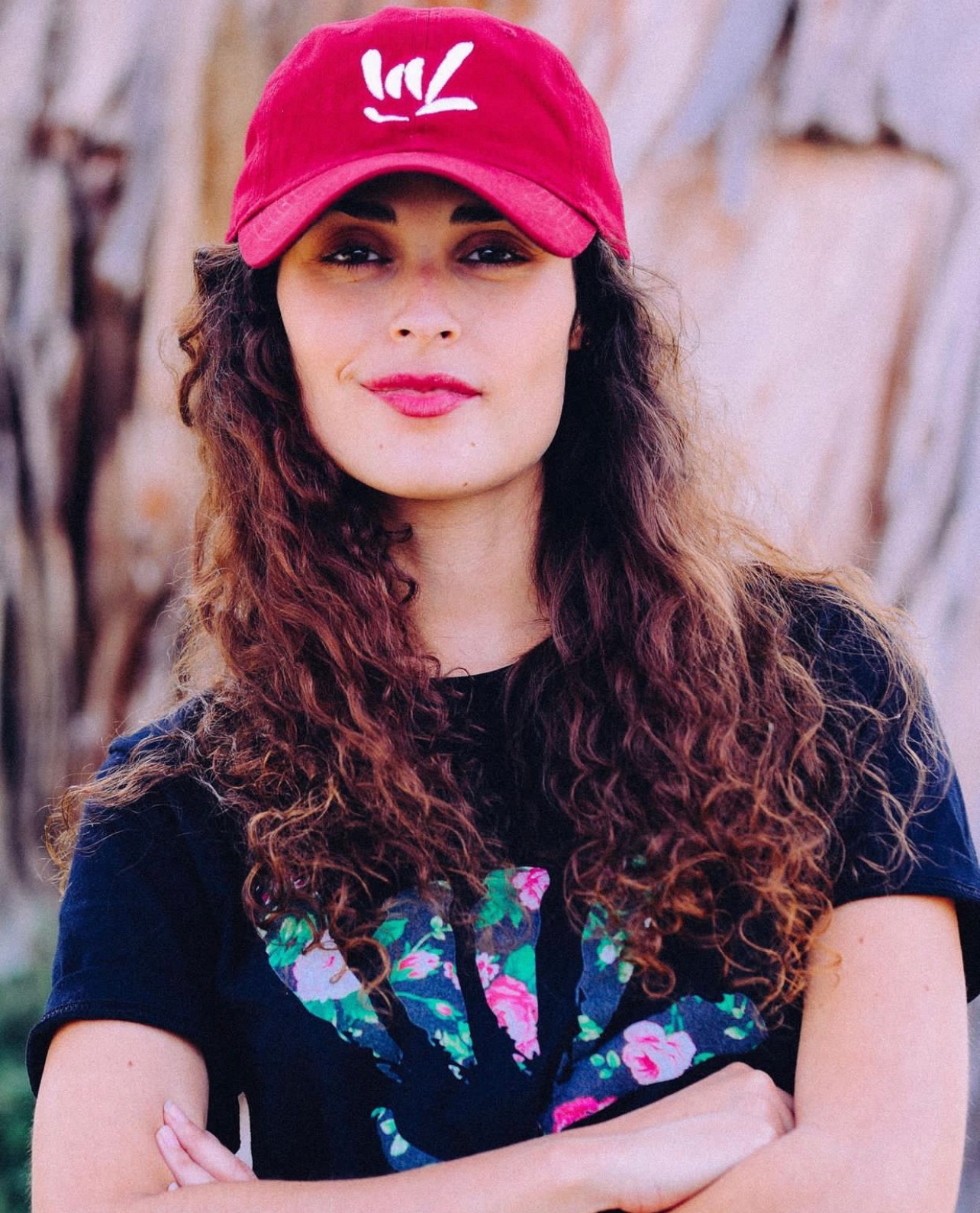 Woman with curly hair wearing a red cap and black T-shirt, standing outdoors with trees in background.