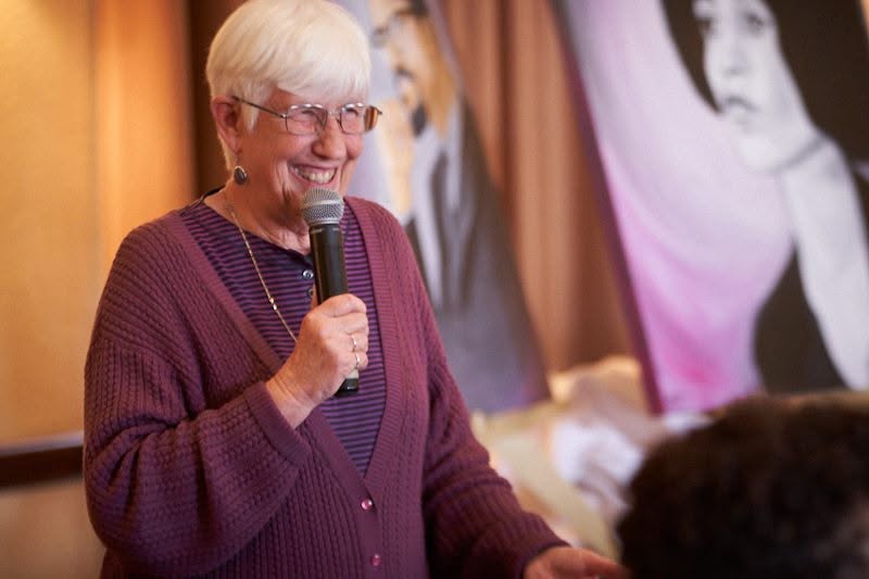 Older woman with glasses smiling and speaking into a microphone, wearing a purple sweater, in a room with a large black-and-white photo in the background.