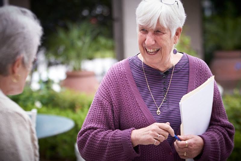 Older woman with short white hair smiling, holding a notebook, talking to a person outdoors in a garden setting.