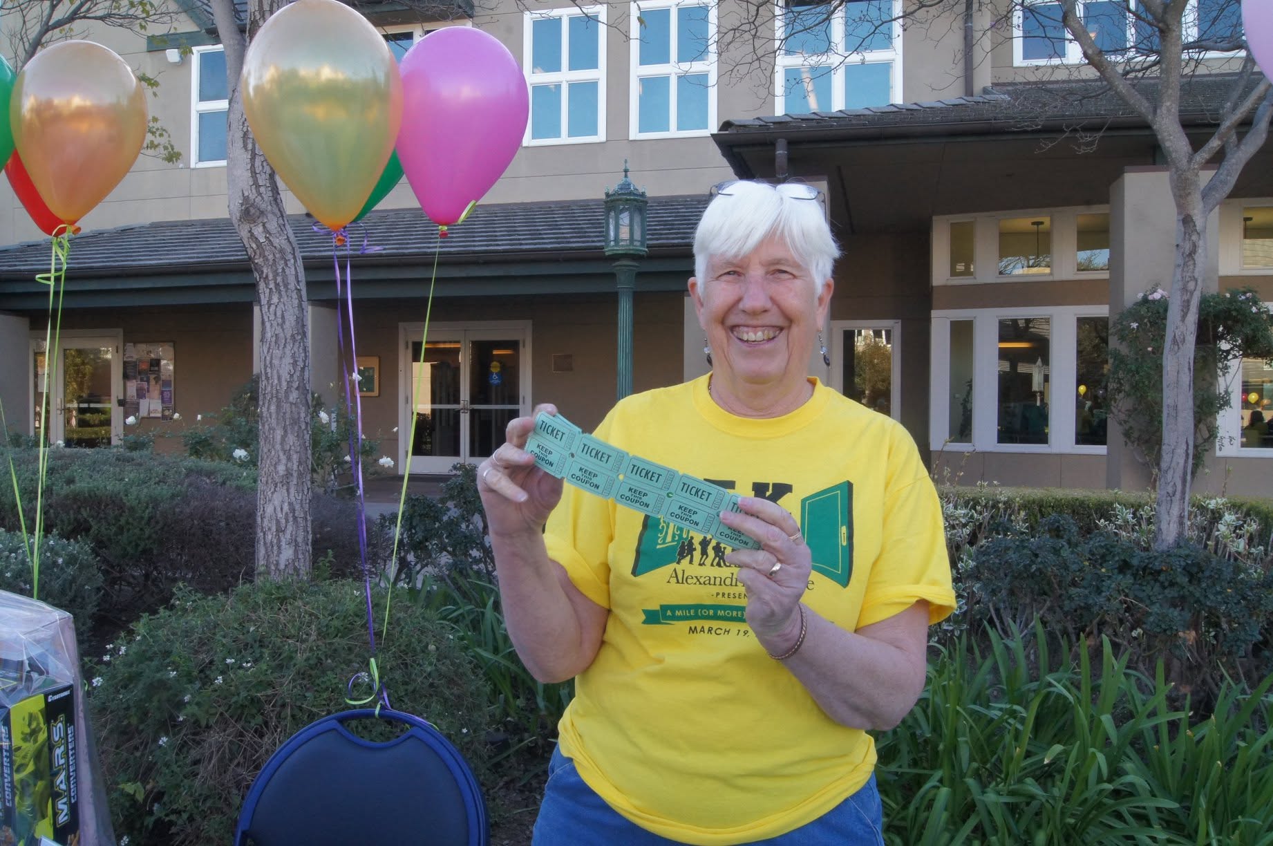 Smiling person with white hair holding a ticket outdoors, with colorful balloons and a building in the background.