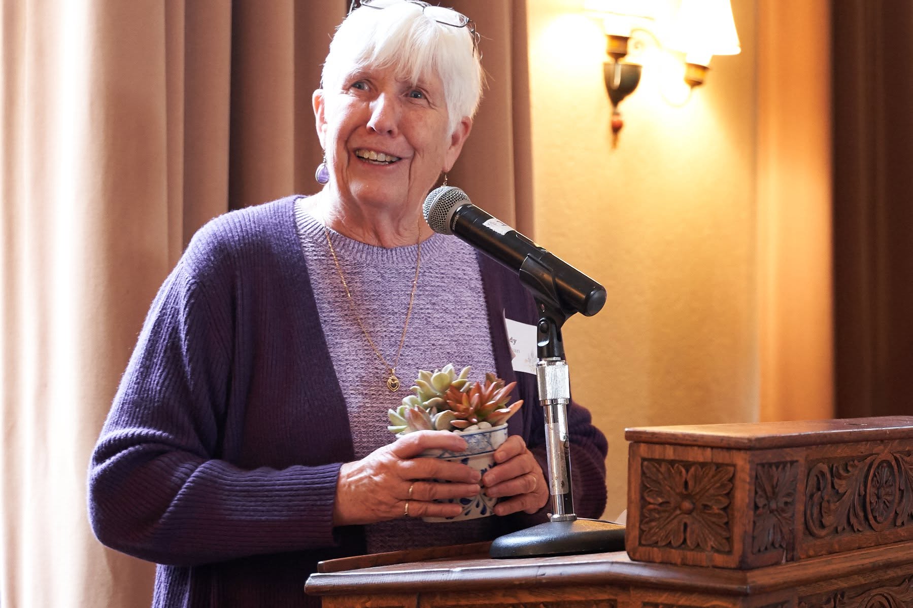 Older woman with white hair speaking into a microphone, holding a small potted plant, standing behind a wooden podium.