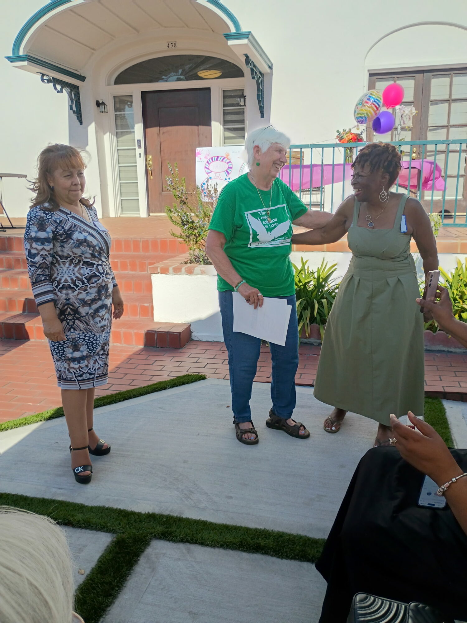 Three women standing outdoors near a small building, with balloons and decorations in the background, engaged in conversation.