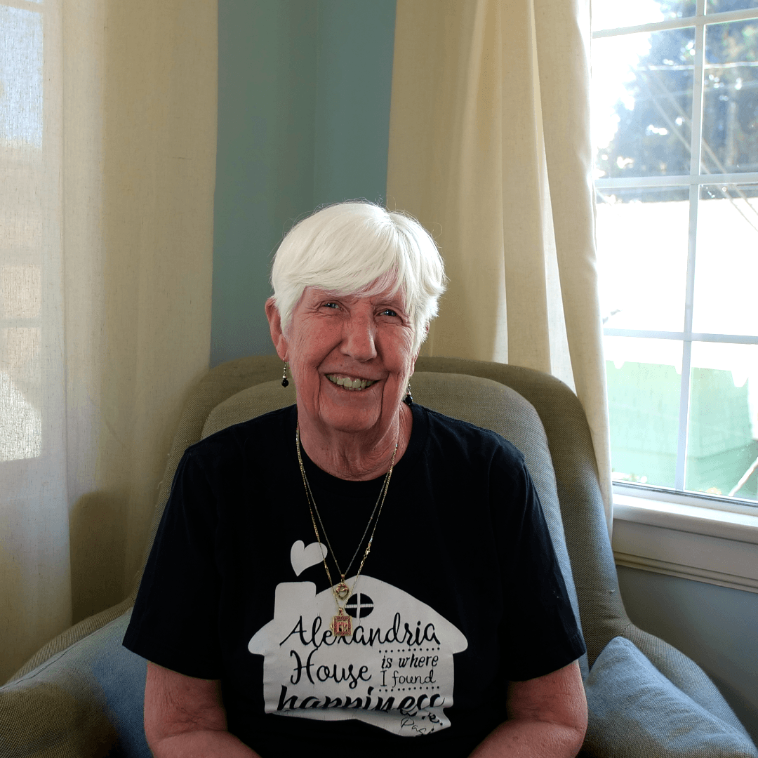 Older woman with short white hair smiling, sitting in a chair near a window with curtains, wearing a black T-shirt with text.