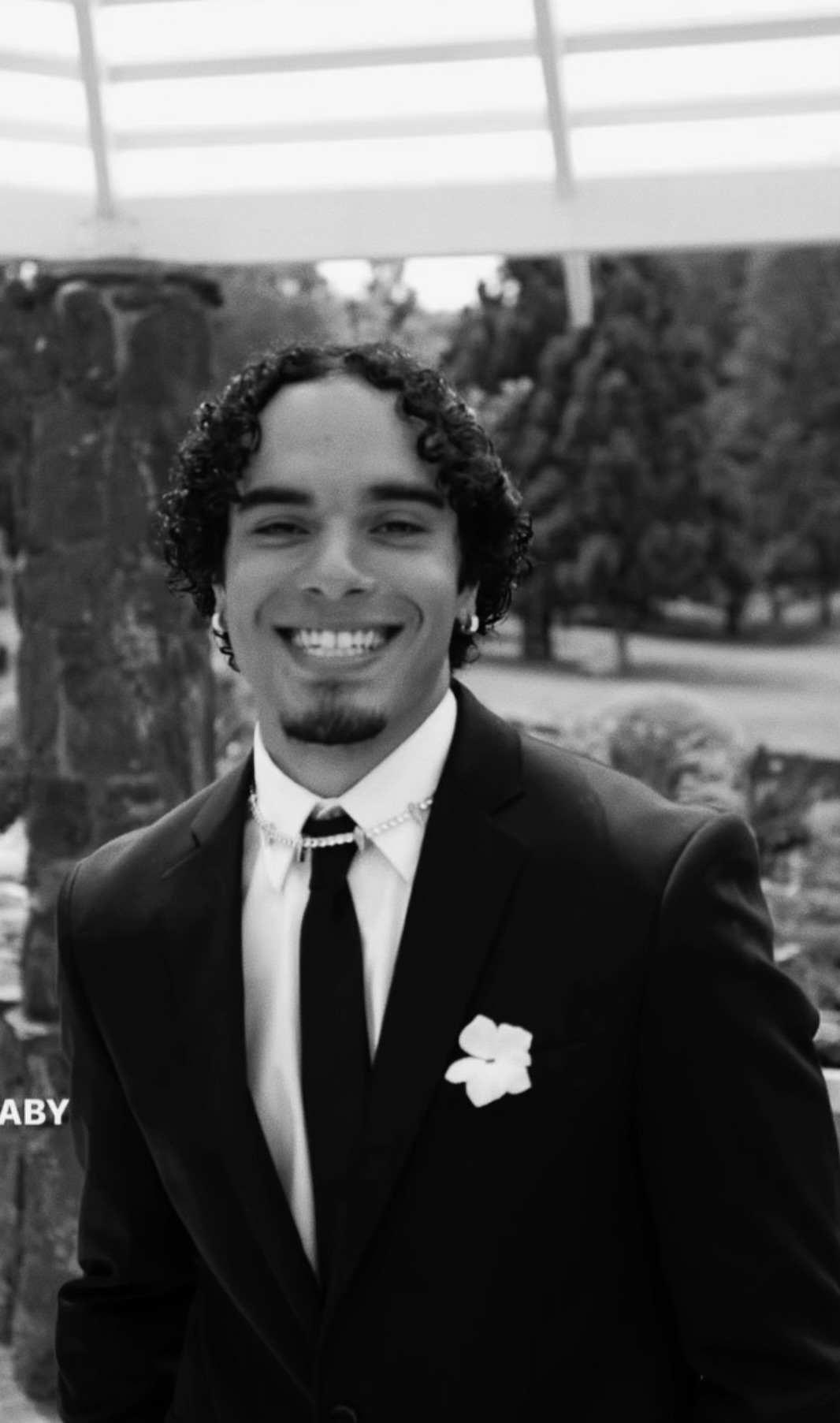 Young man in a suit smiling outdoors, wearing a tie and a flower on his lapel, with trees in the background.