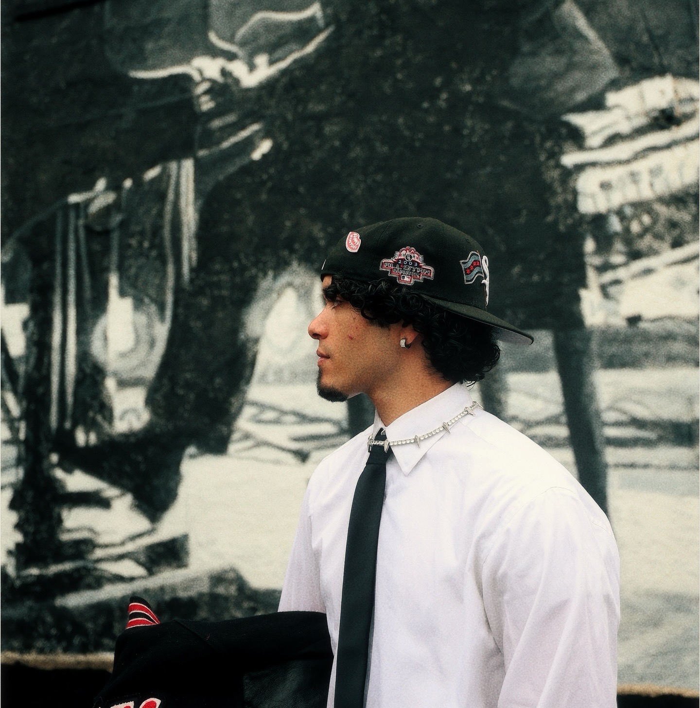 Young man in white shirt, black tie, and black cap with patches, standing sideways outdoors, with a black and white mural background.