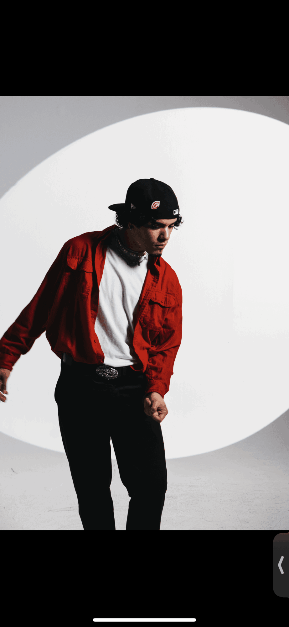 Person wearing a red jacket, white shirt, black pants, and a black cap, standing against a white background with a circular design.