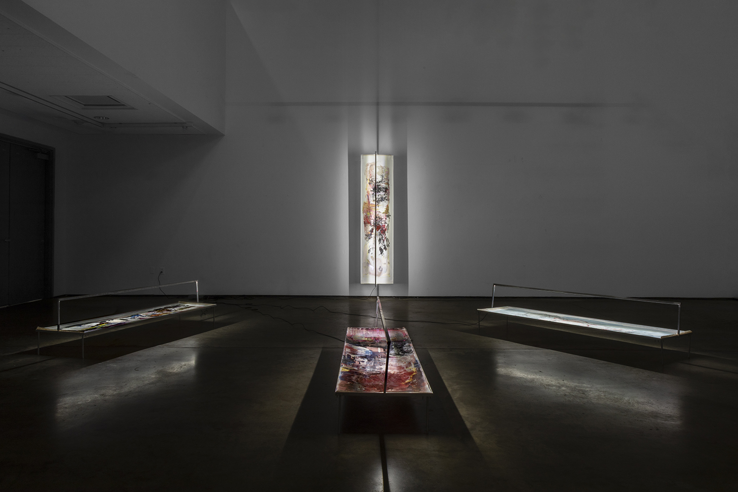 A narrow vertical window with light shining through, flanked by two display cases in a dark room.