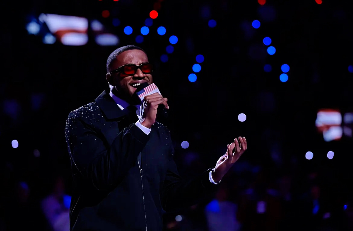 Person singing into a microphone on stage with a dark background and colorful lights, smiling, wearing glasses and a dark outfit.