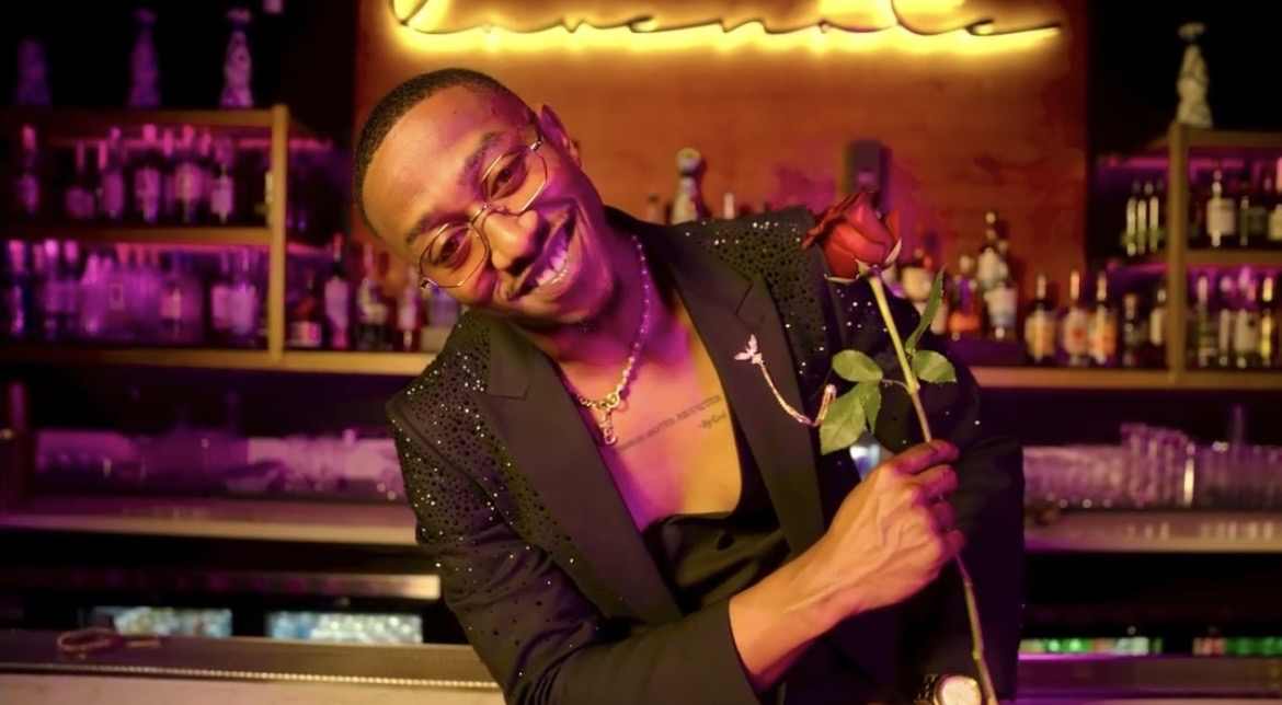 Person smiling and holding a flower in a bar with colorful lighting and bottles in the background.