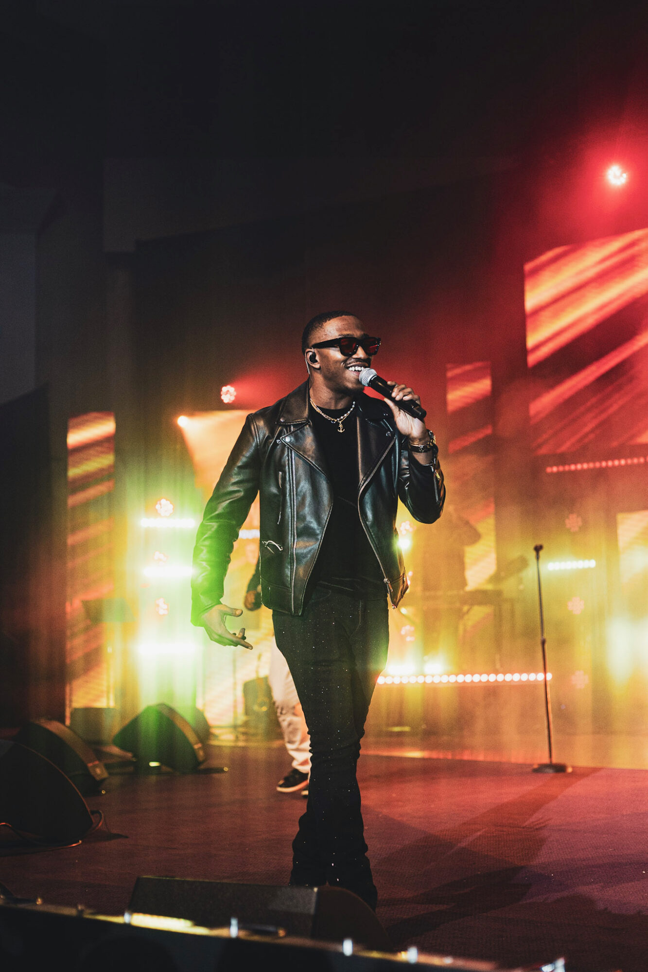 Male performer singing into a microphone on stage with colorful lights, wearing sunglasses and a leather jacket.