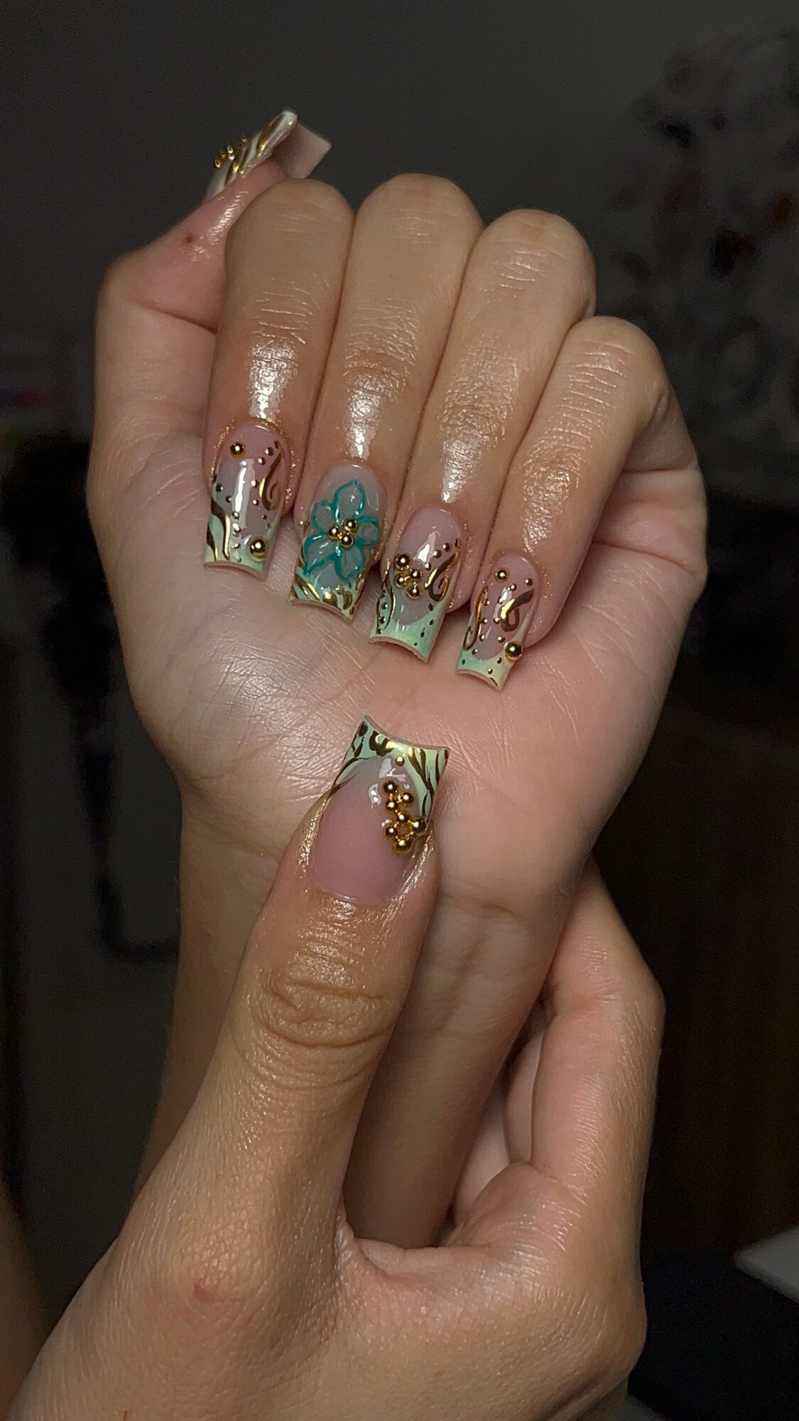 Close-up of a hand with decorated nails featuring gold accents, blue and green designs, and rhinestones.