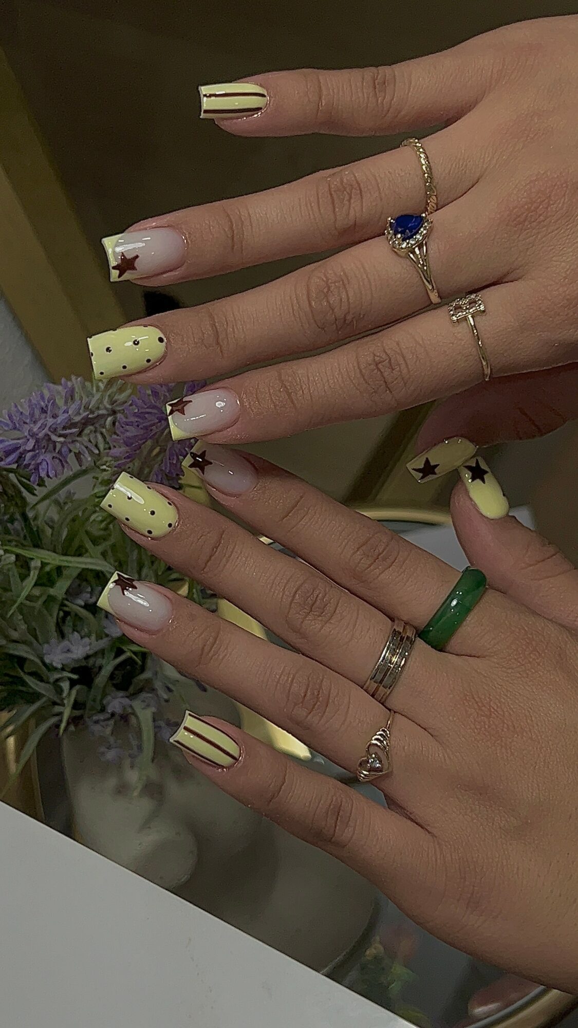 Two hands with decorated nails and rings, holding a purple flower, with a blurred background.