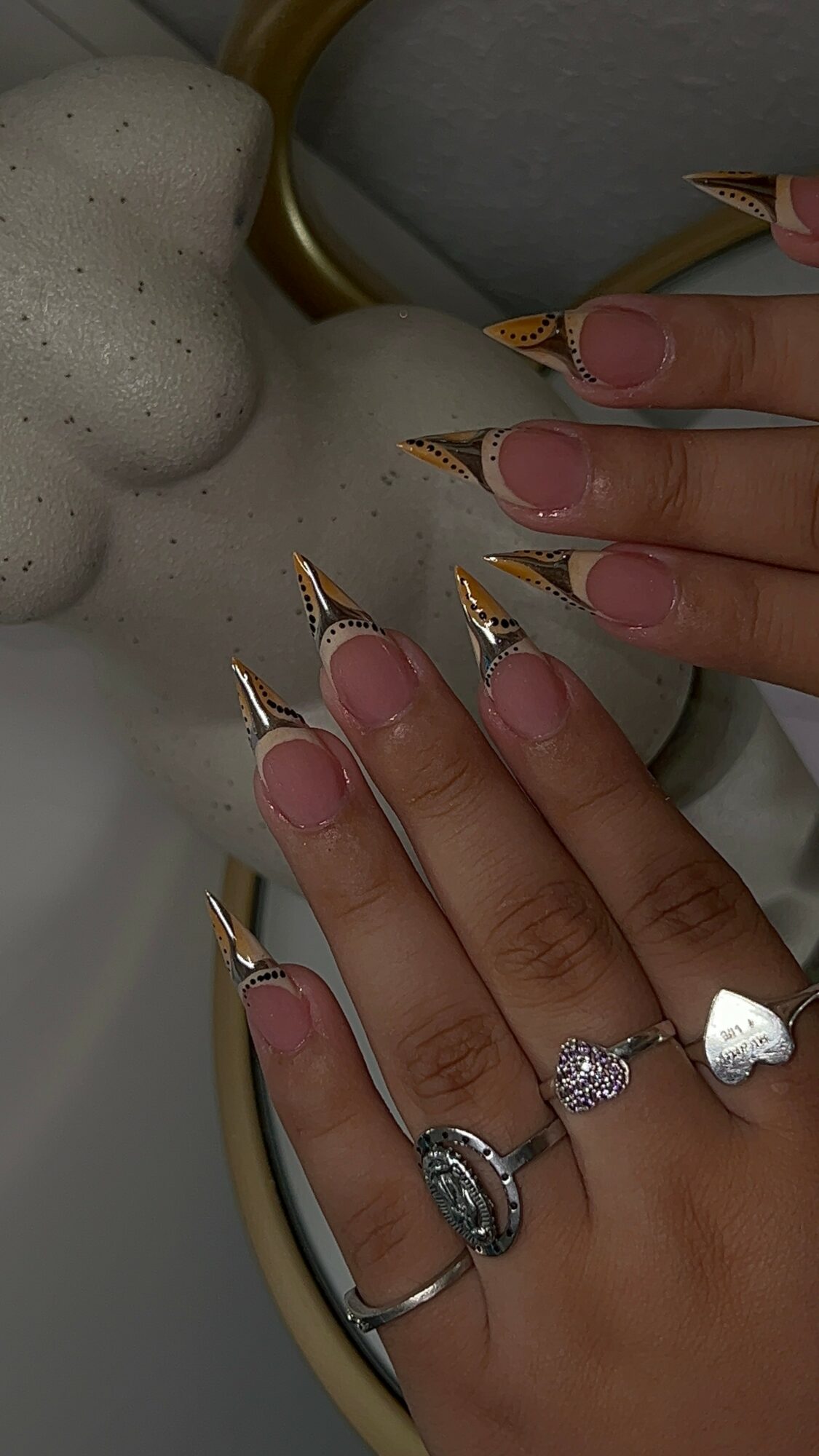 Hand with long, decorated nails and rings, resting on a textured surface with a gold-colored object nearby.