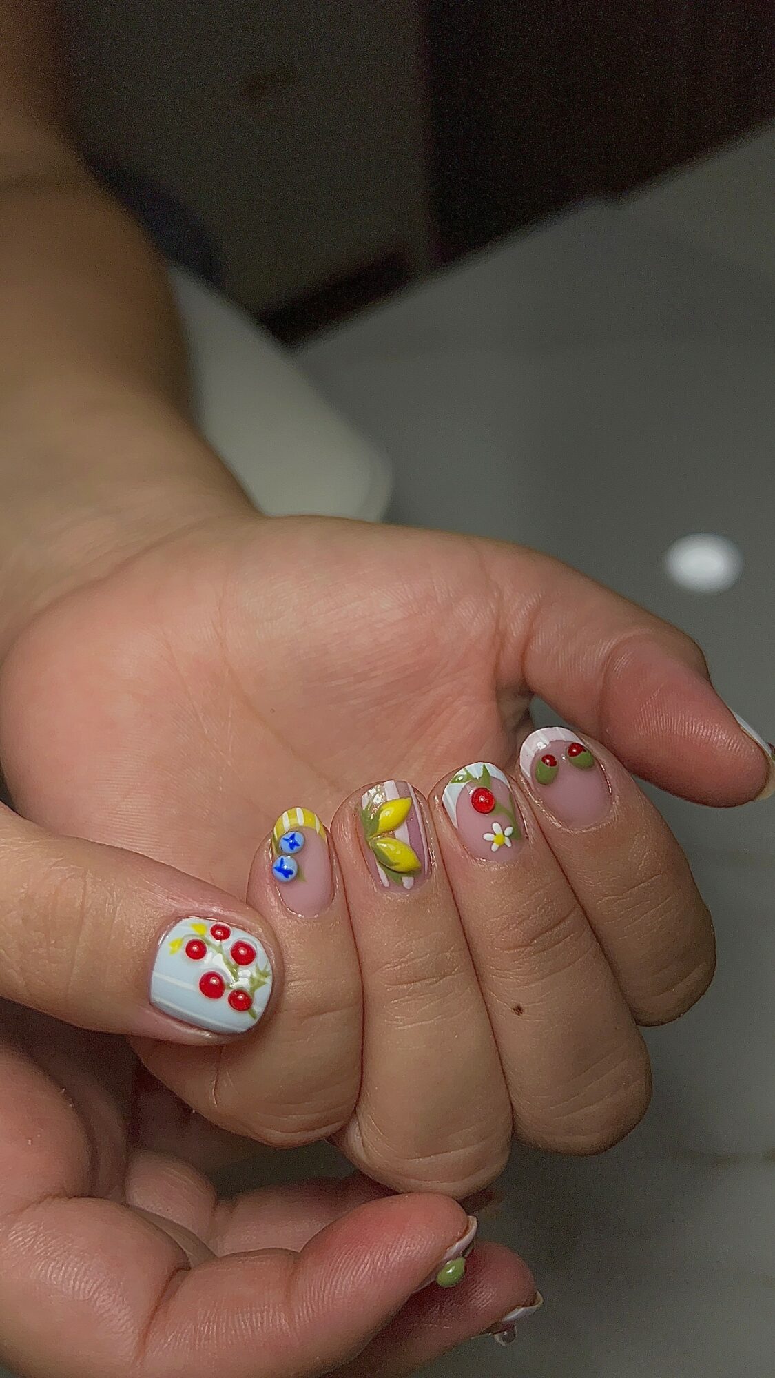 Hand with decorated nails featuring colorful floral and abstract designs, some with rhinestones, on a white background.