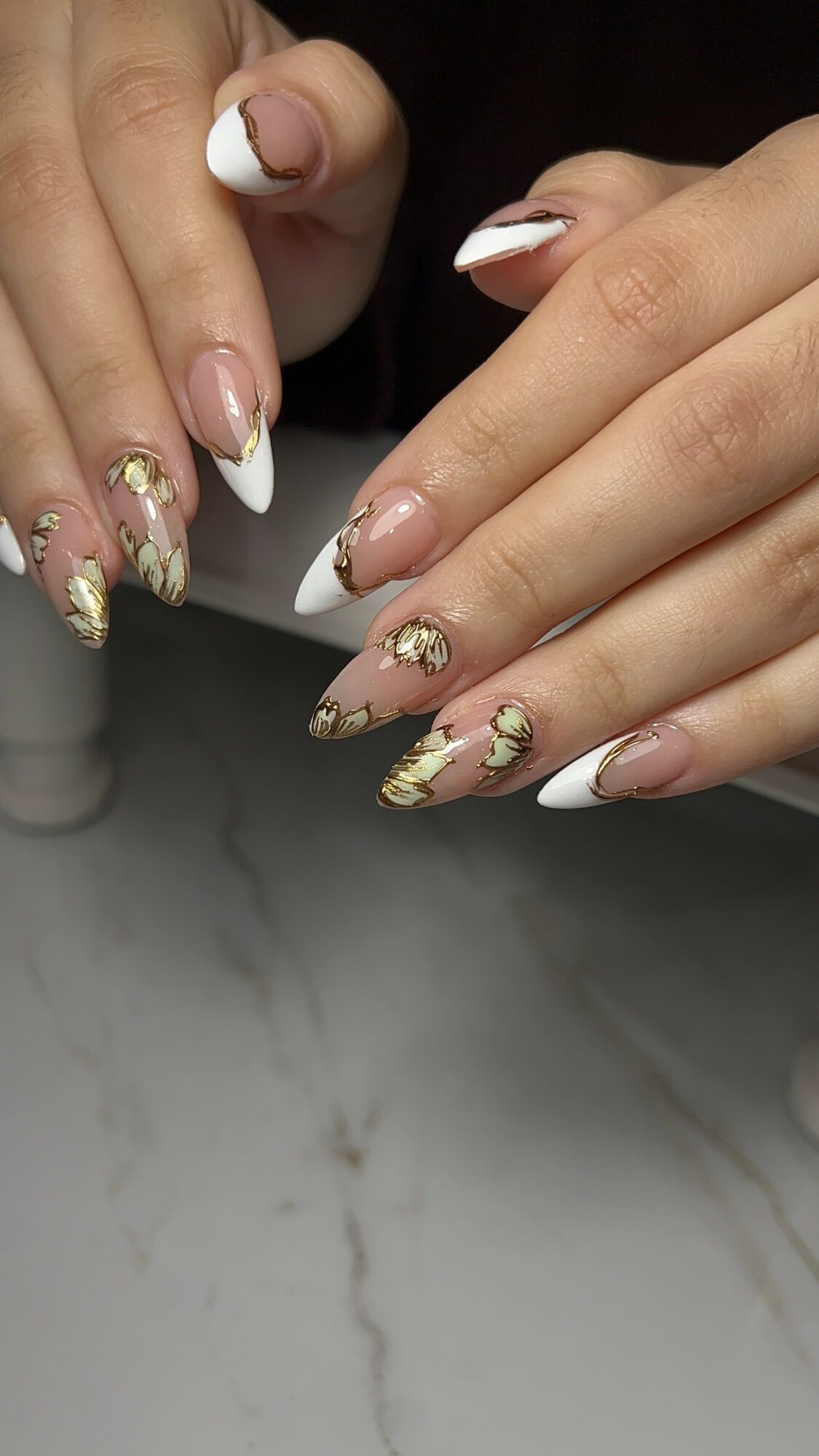 Hands with decorated nails, some with white tips and gold accents, resting on a marble surface.