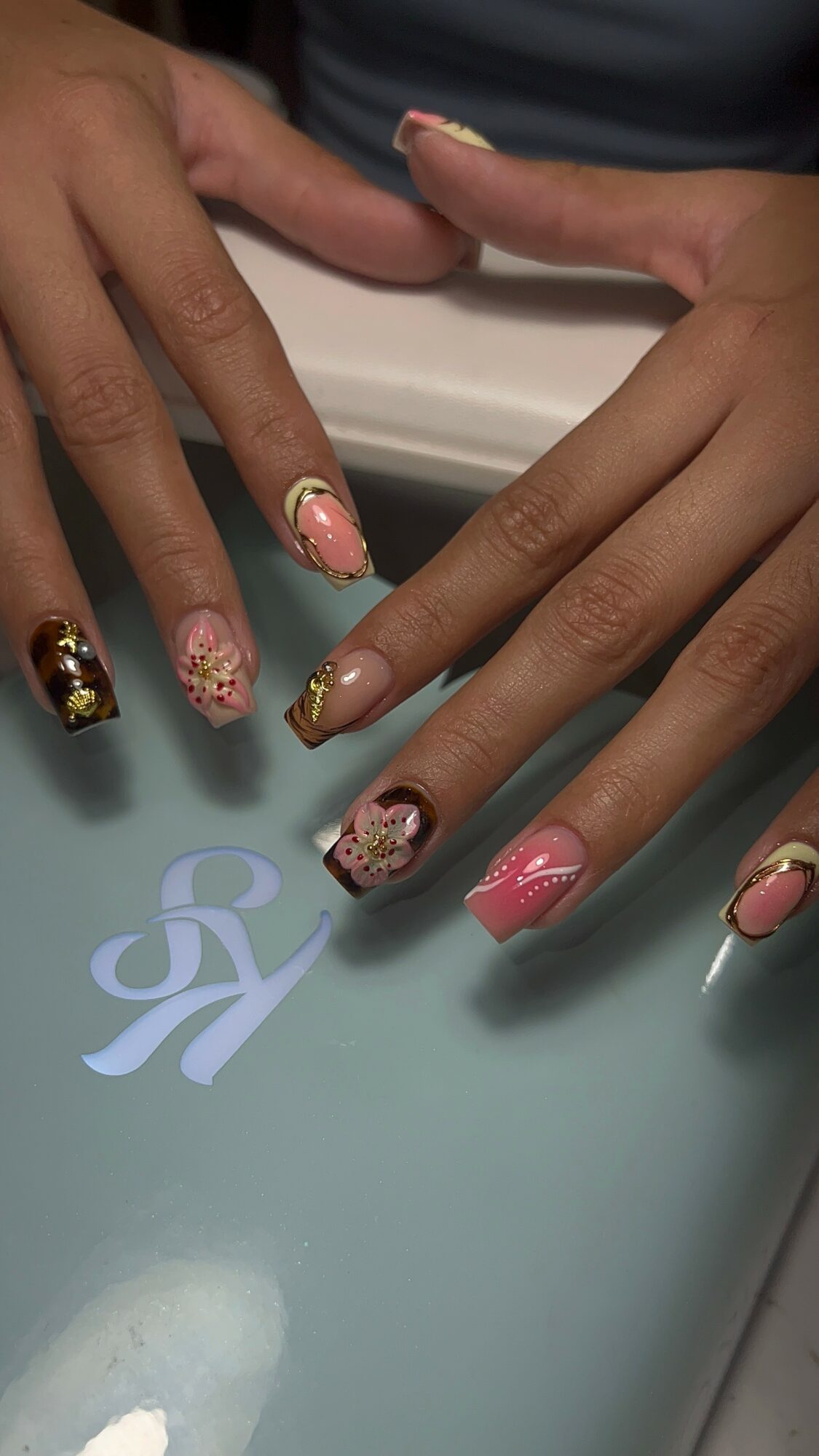 Two hands with decorated nails resting on a surface with a logo, nails painted with various designs and colors.