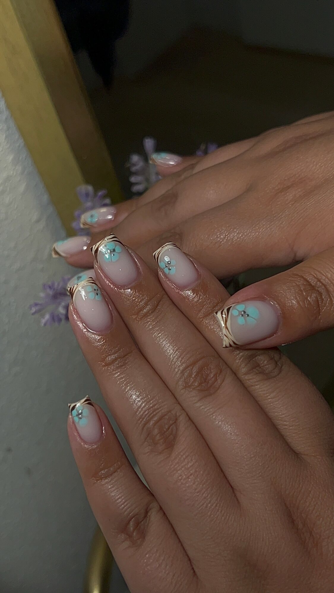 Hands with decorated nails resting on a surface, nails have light blue polish with floral designs and rhinestones.