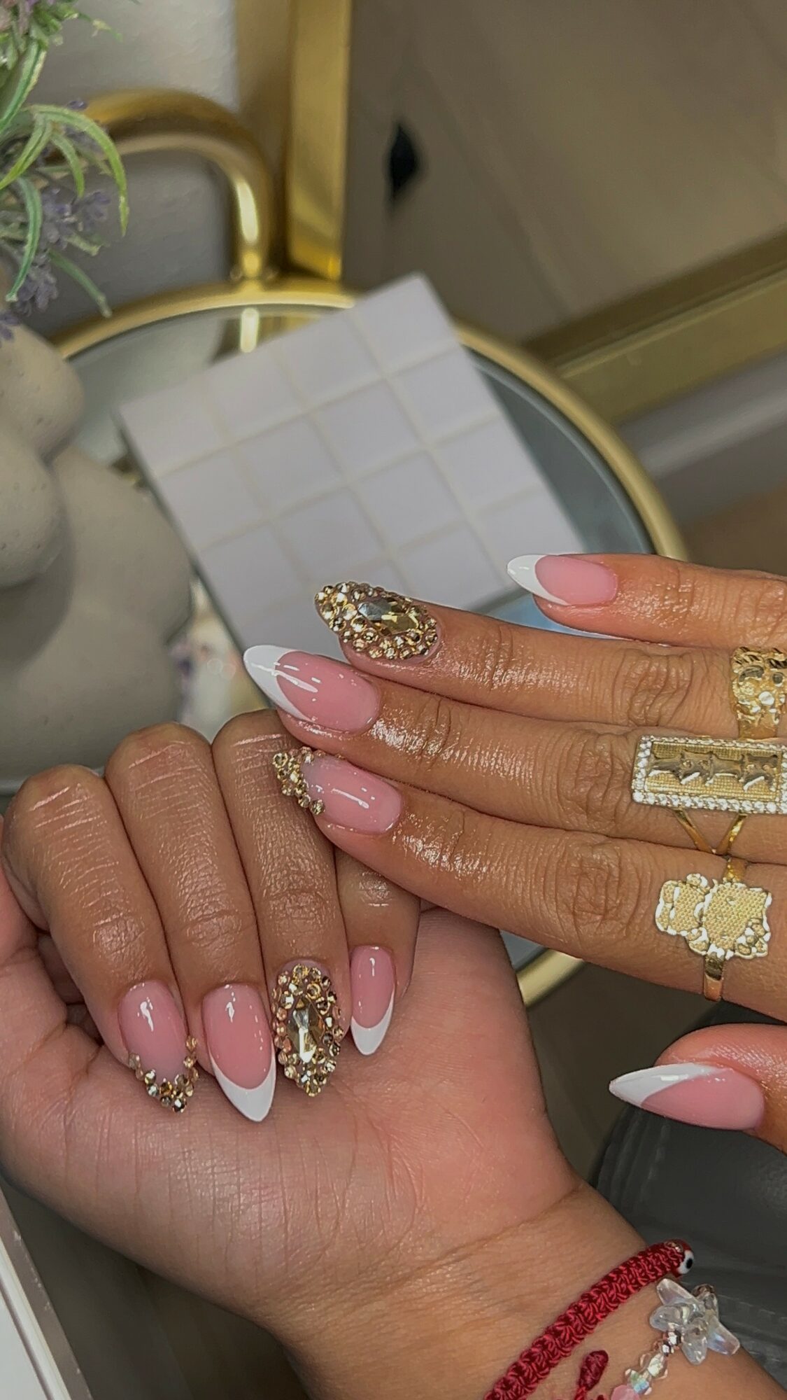 Two hands with manicured nails and decorated rings, one hand resting on a surface with a white tiled background.