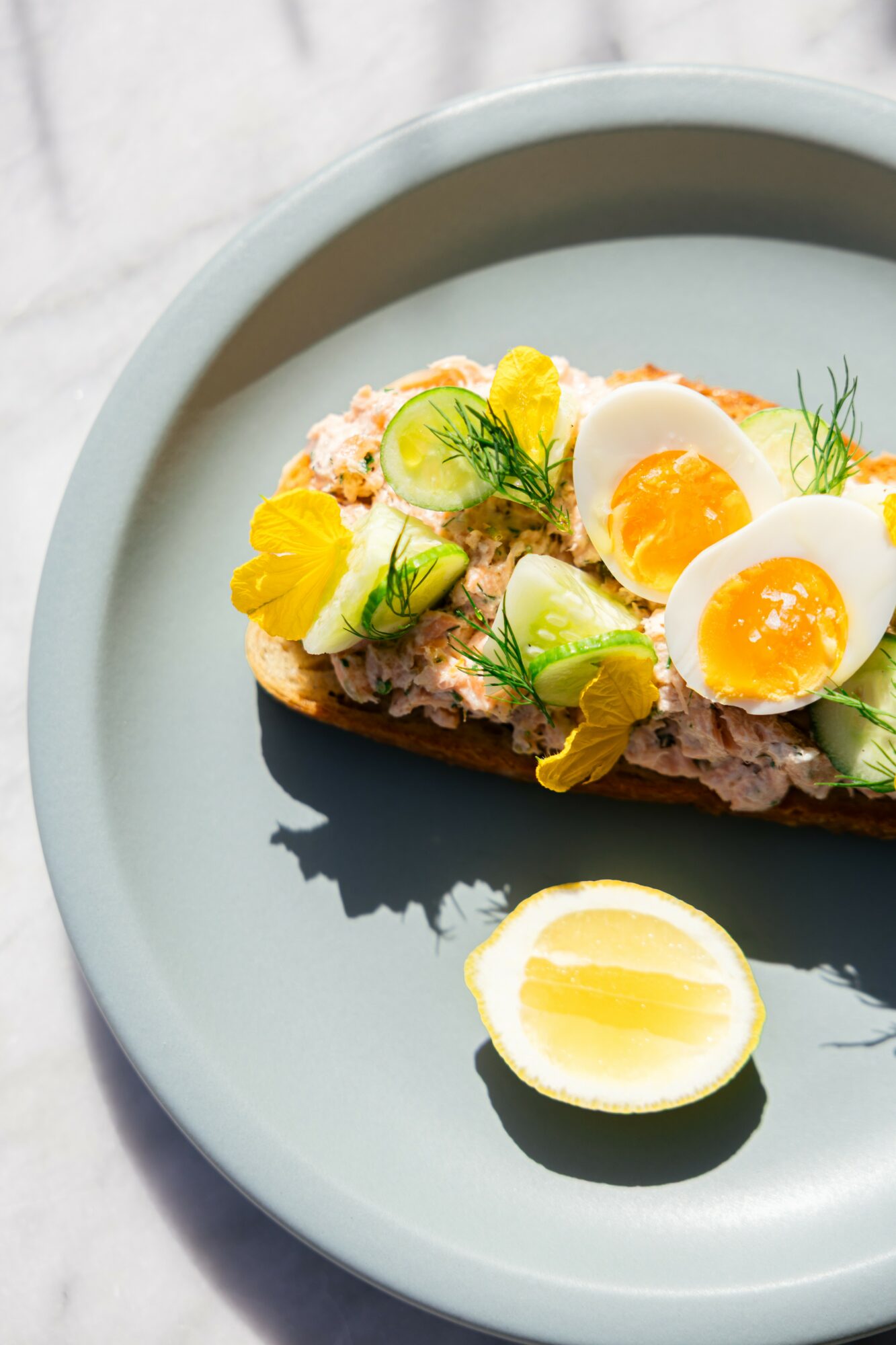 Open-faced sandwich with halved soft-boiled eggs, lemon wedge, herbs, and yellow edible flowers on a blue plate.