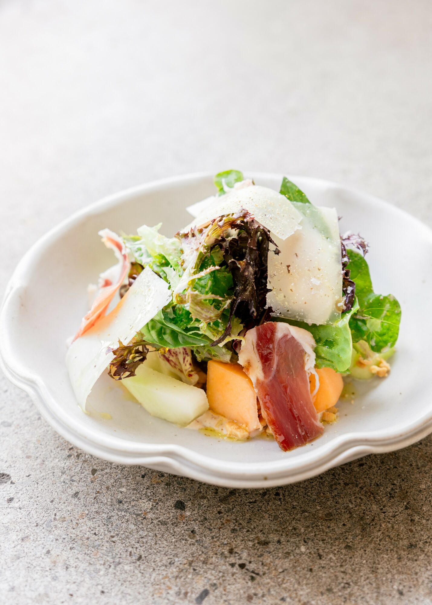 A small dish with a layered salad including greens, cheese, and slices of meat, on a gray surface.