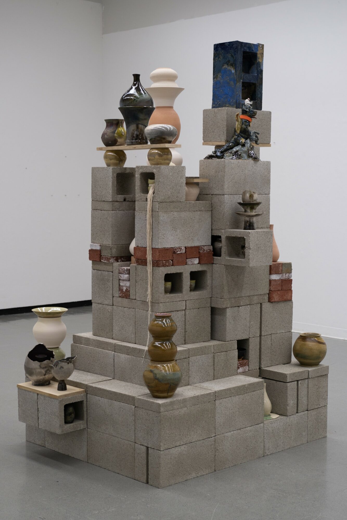 Sculpture composed of stacked concrete blocks with various vases and objects on top, displayed in a gallery setting.