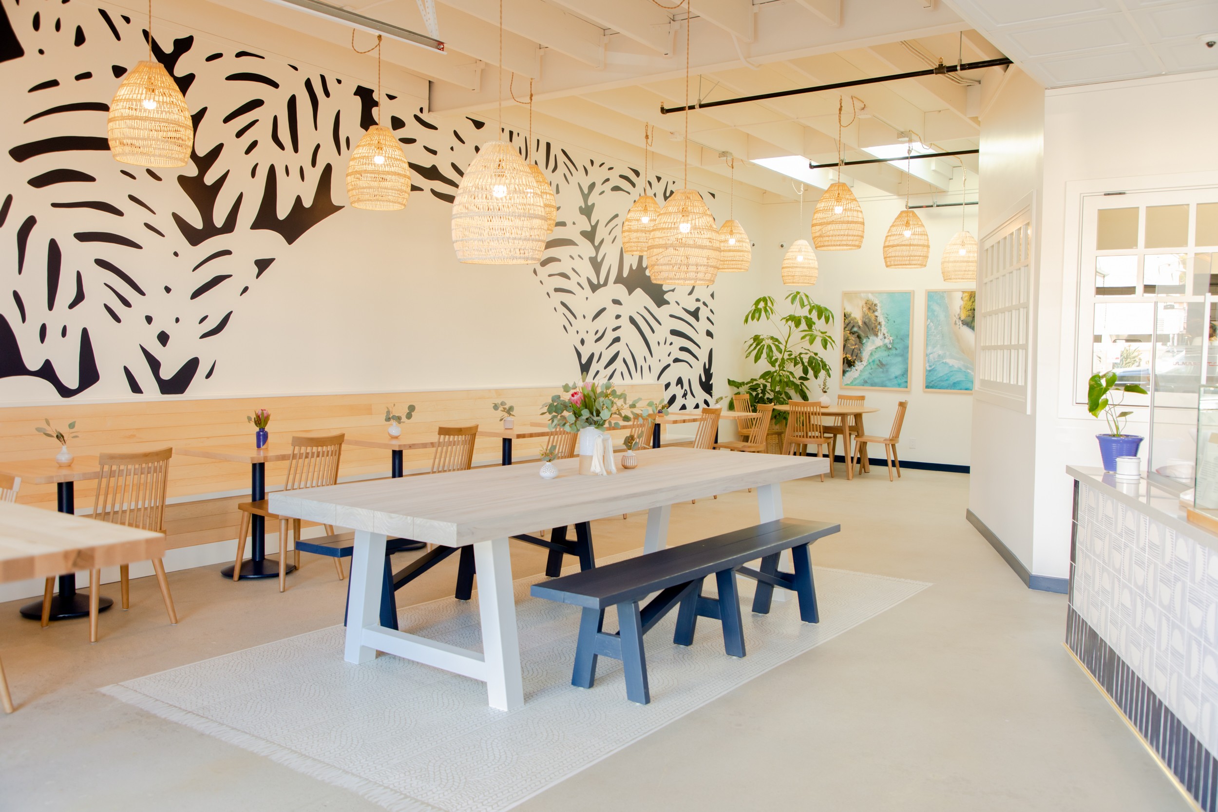 Bright dining area with a large white table, wooden chairs, hanging pendant lights, and a decorative wall mural with tropical leaves.