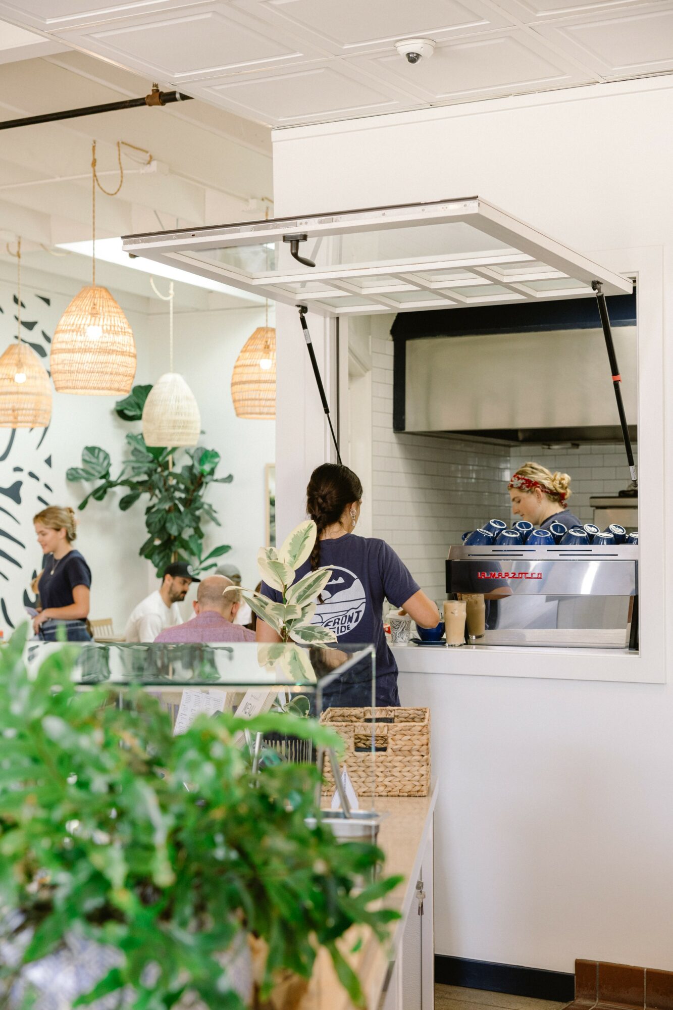 People working behind a counter in a cafe with hanging lights and plants, viewed from outside through a window.