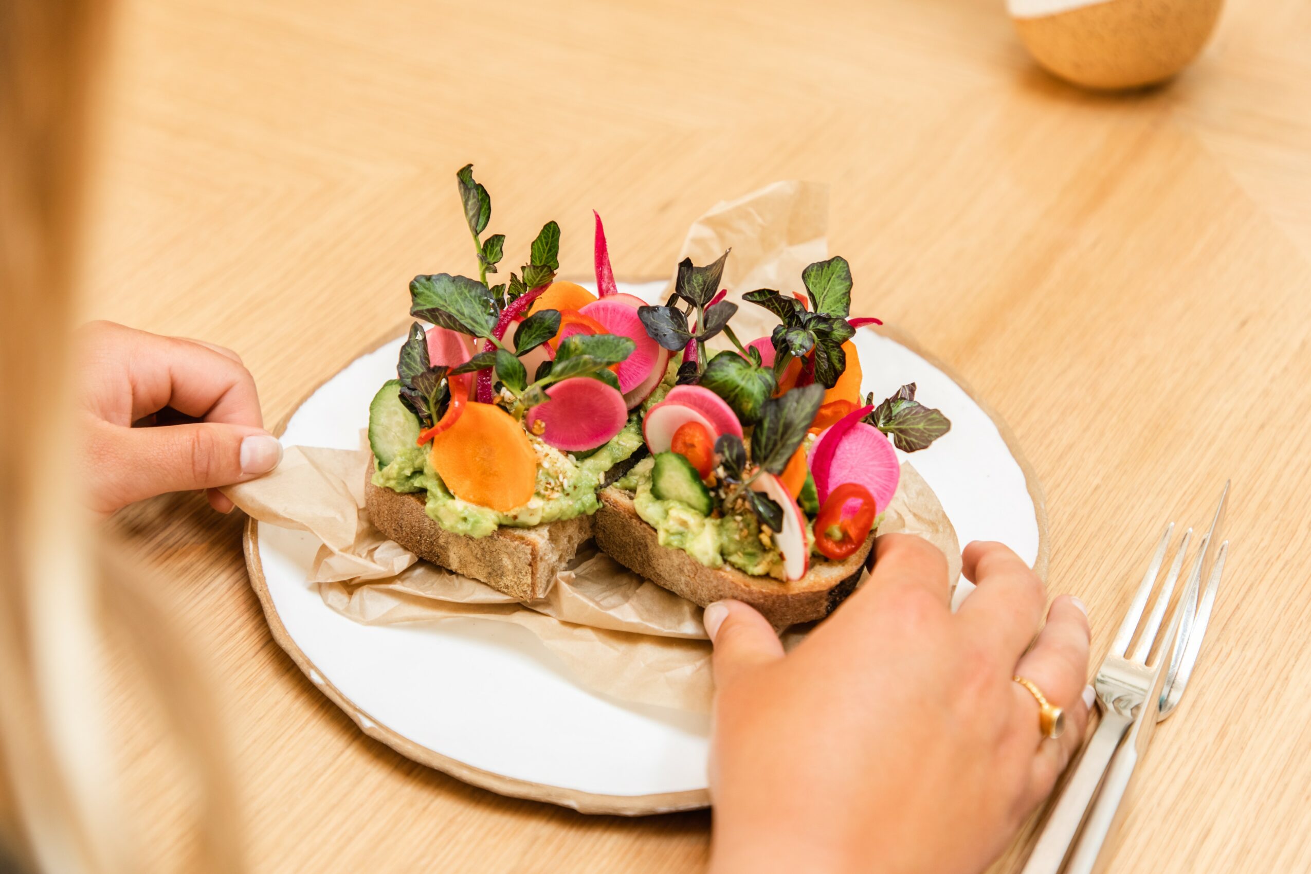 Person holding a plate with three open-faced sandwiches topped with vegetables and herbs, on a wooden table.