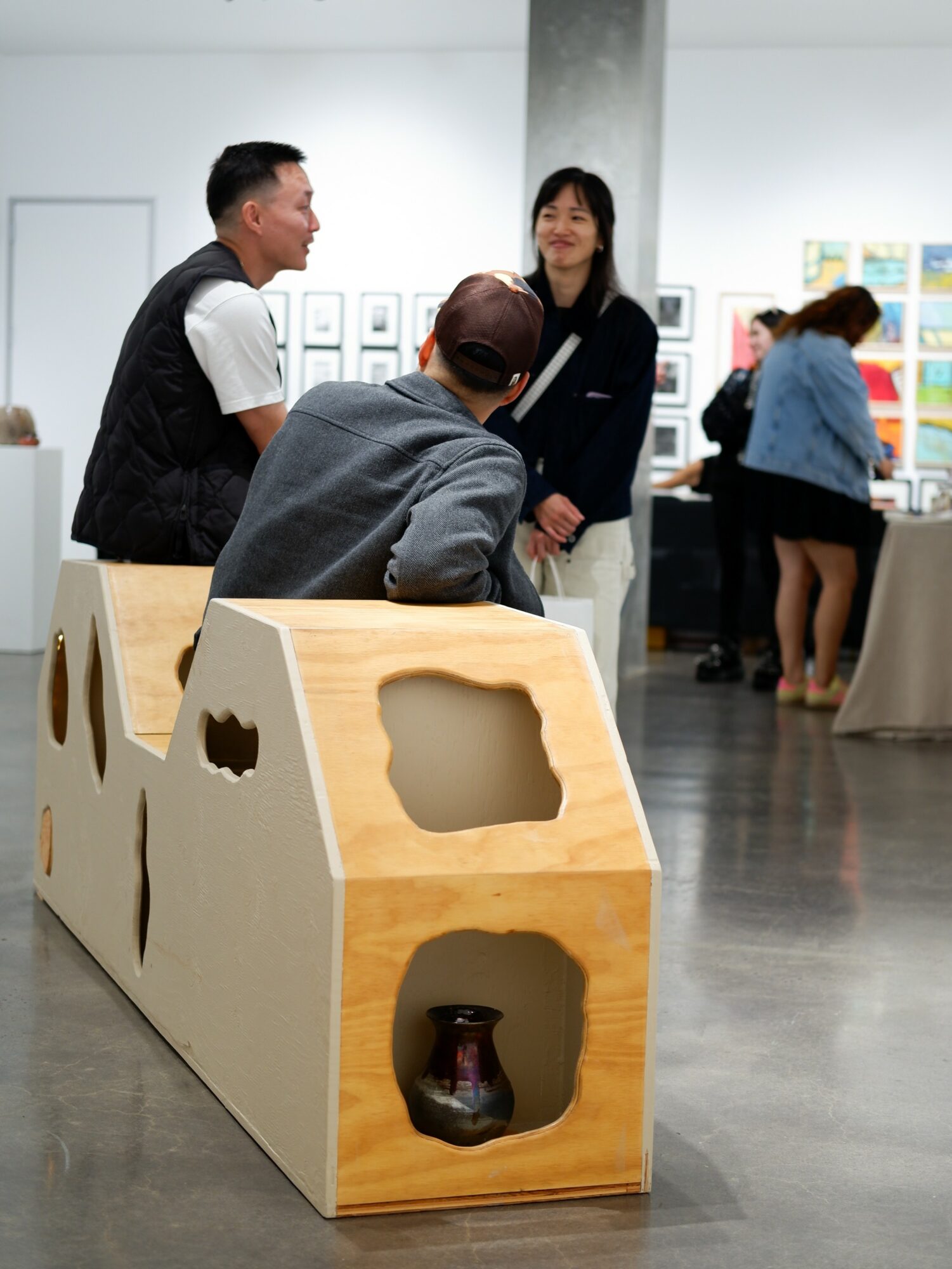 Three people engaged in conversation in an art gallery, with artwork displayed on the walls and a wooden display in the foreground.