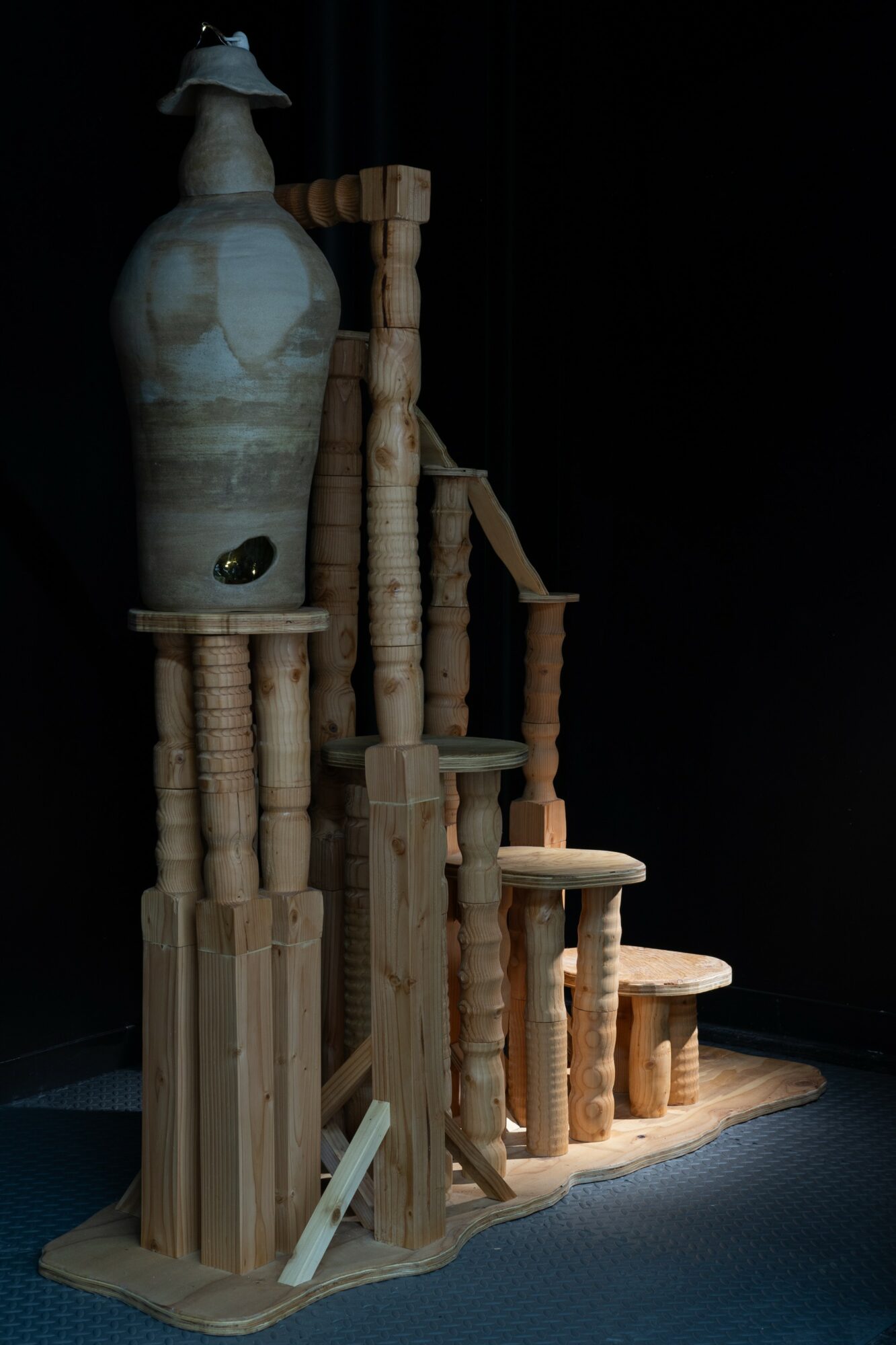 Wooden sculpture with a large ceramic vase on top, set against a dark background, with various wooden columns and platforms.