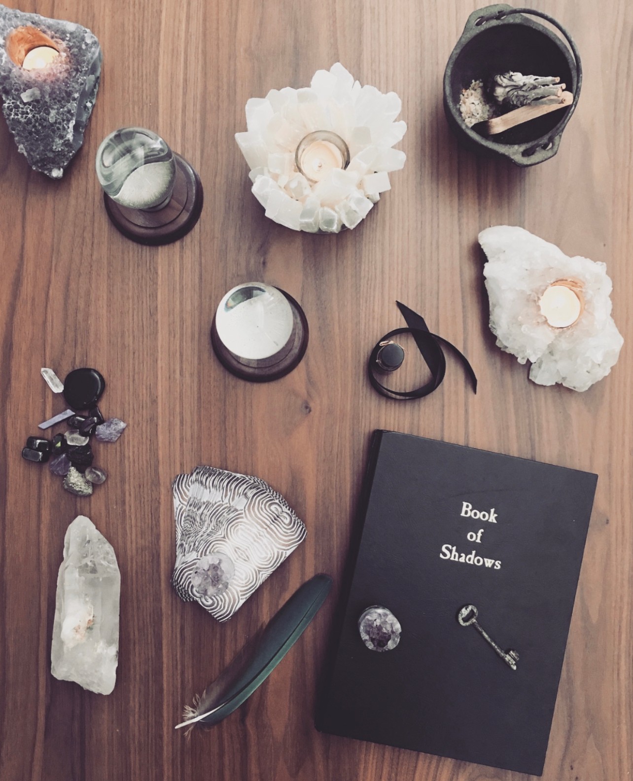 Flat lay of crystals, candles, a book titled 'Book of Shadows', and decorative objects on wooden surface.