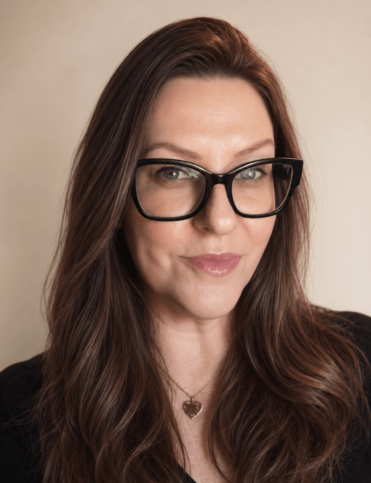 Woman with long brown hair wearing glasses and a necklace, looking at the camera, neutral background.