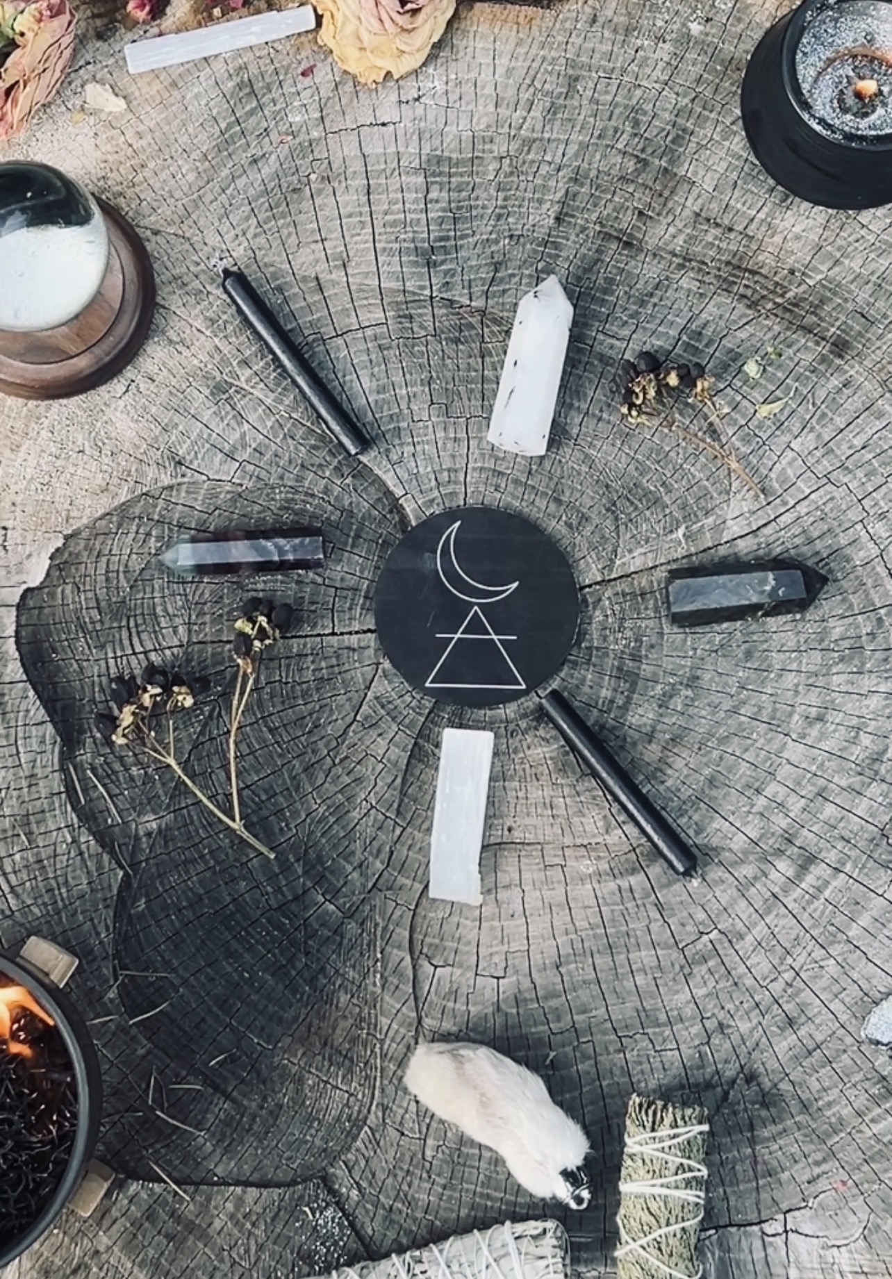 A circular arrangement of candles, crystals, and decorative objects on a wooden surface with a black central piece featuring a moon and triangle symbol.