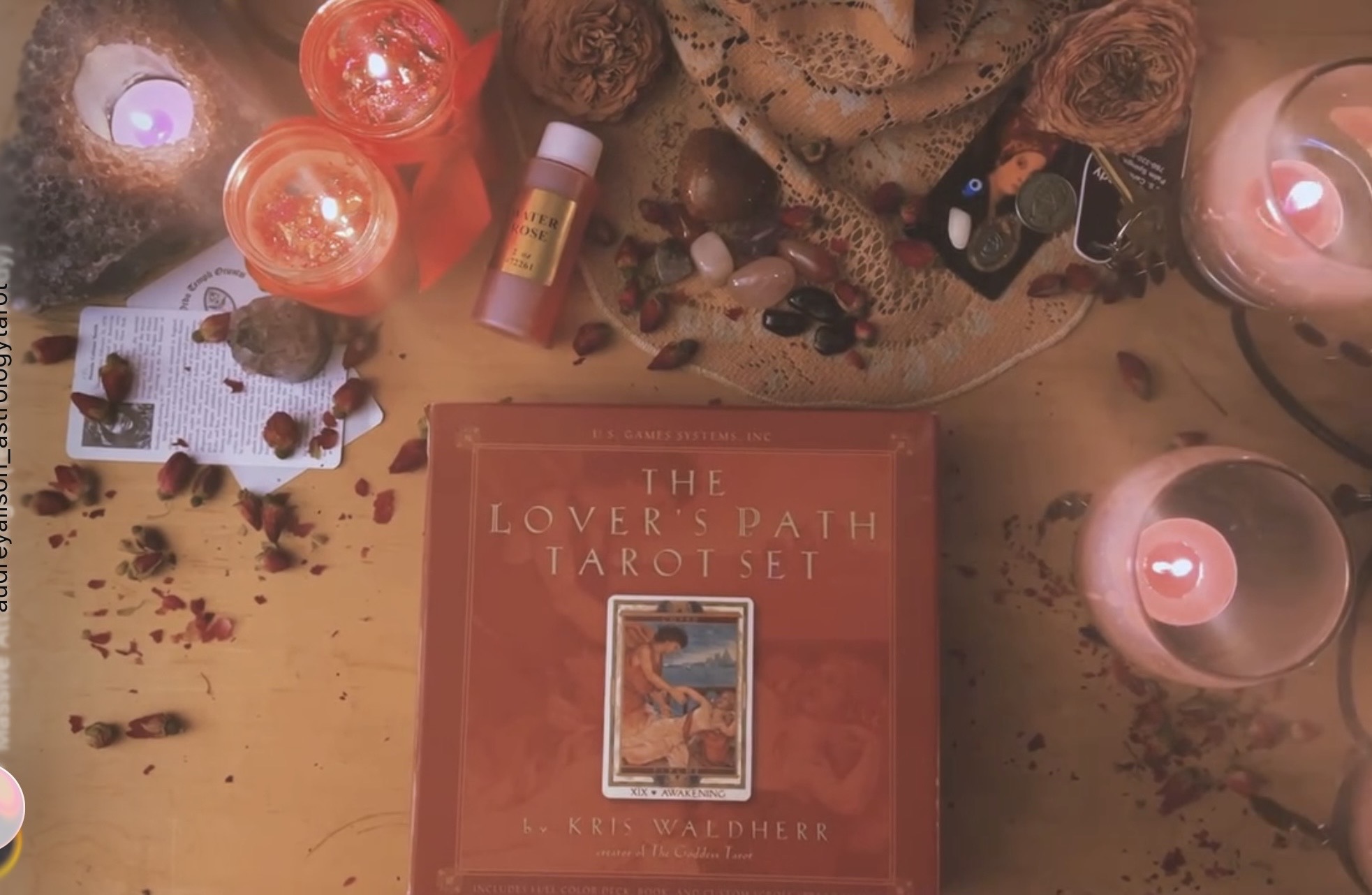 A table with candles, a book titled 'The Lover's Path Tarot Set,' crystals, and various small objects, illuminated by warm lighting.
