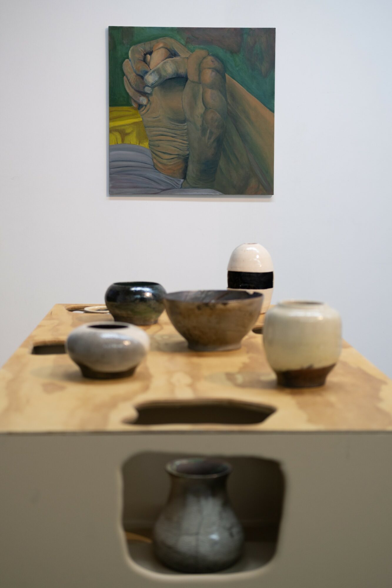 Painting of two hands intertwined hangs above a wooden table with ceramic bowls and a vase.